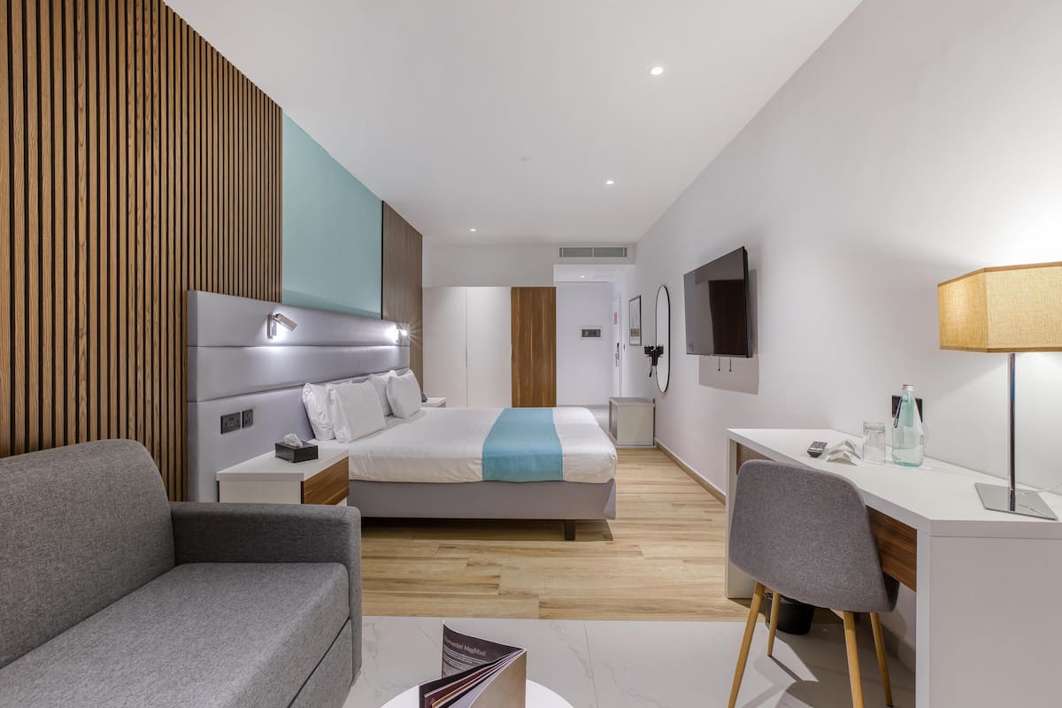 Quarto de hotel moderno com cama, escrivaninha, cadeira e sofá.