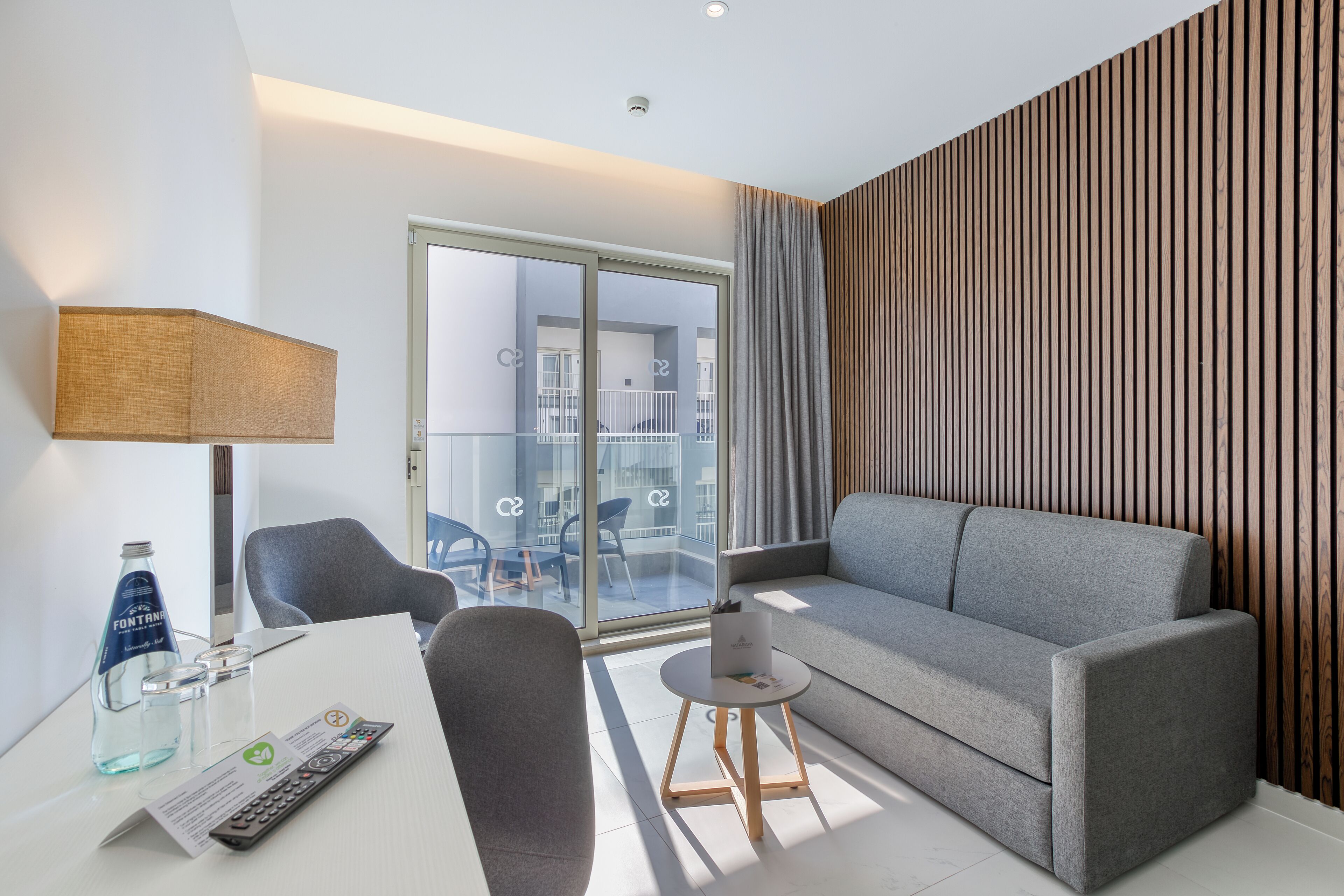 A modern hotel room with a grey sofa, a small round table with a lamp, a glass of water, and a remote control.