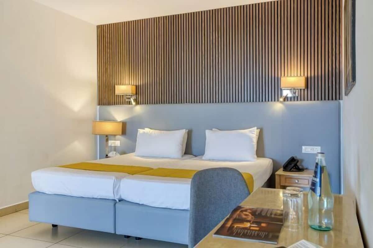 Quarto de hotel com cama, mesas de cabeceira, escrivaninha e luminária fixada na parede.