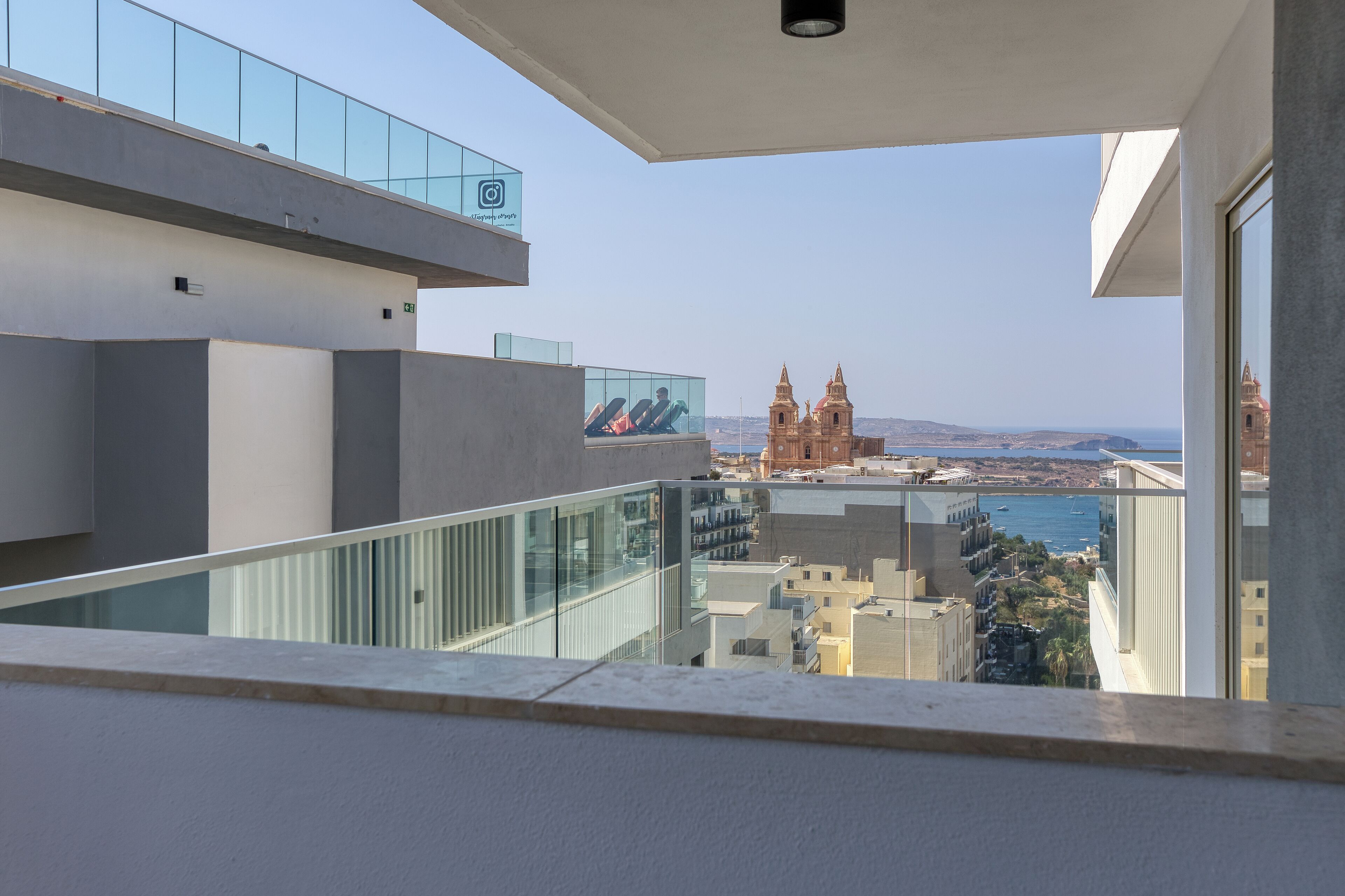 A modern building with a glass balcony overlooking a coastal cityscape, including a prominent church.