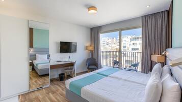 A hotel room with a large bed, a desk, a chair, a TV, and a balcony with a view of buildings.