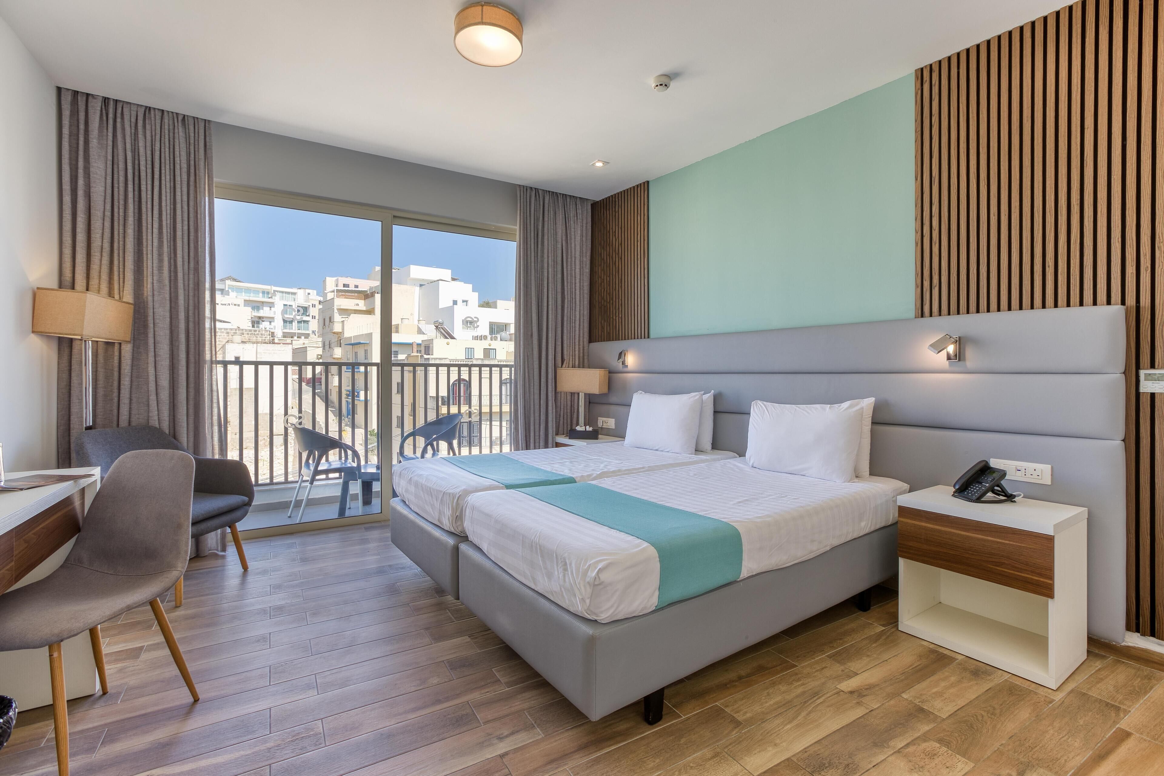 A modern hotel room with a large bed, a desk, and a chair. There is a balcony with a view of the city.