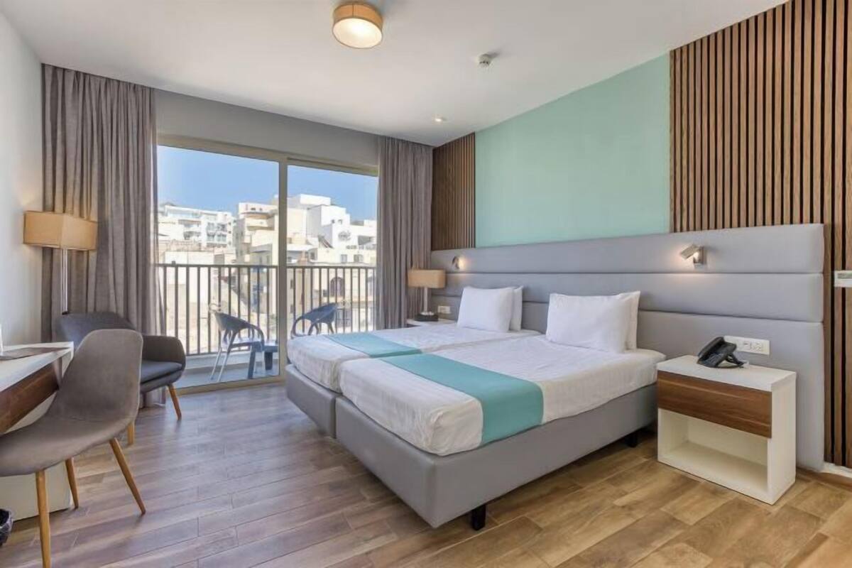 Quarto de hotel com cama, escrivaninha, cadeira, mesa de cabeceira e vista da cidade através de grandes janelas.