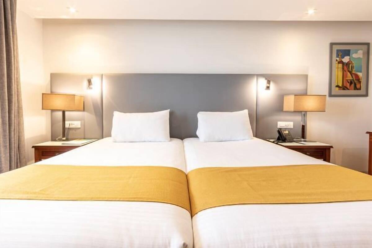 Quarto de hotel com duas camas, cabeceiras, abajures, um quadro na parede e uma cortina.