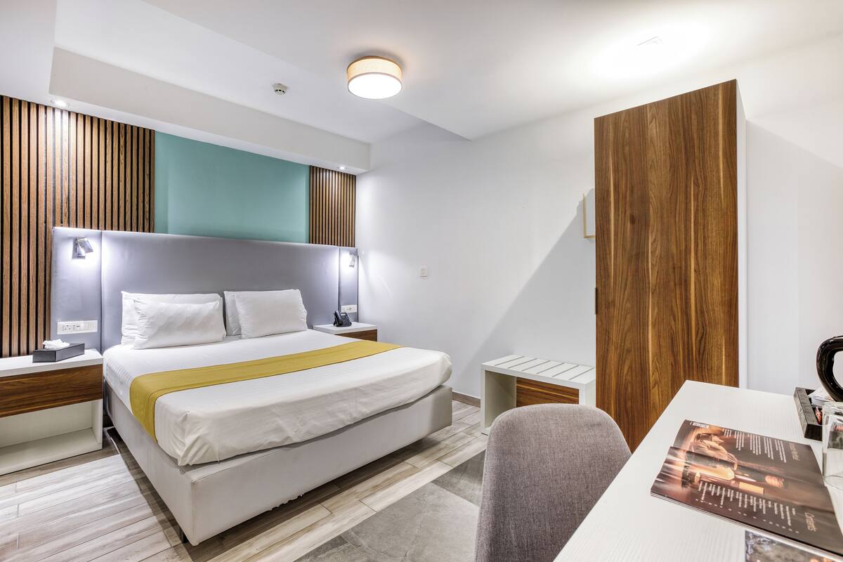 Quarto de hotel com uma cama grande, mesas de cabeceira, escrivaninha e guarda-roupa de madeira.