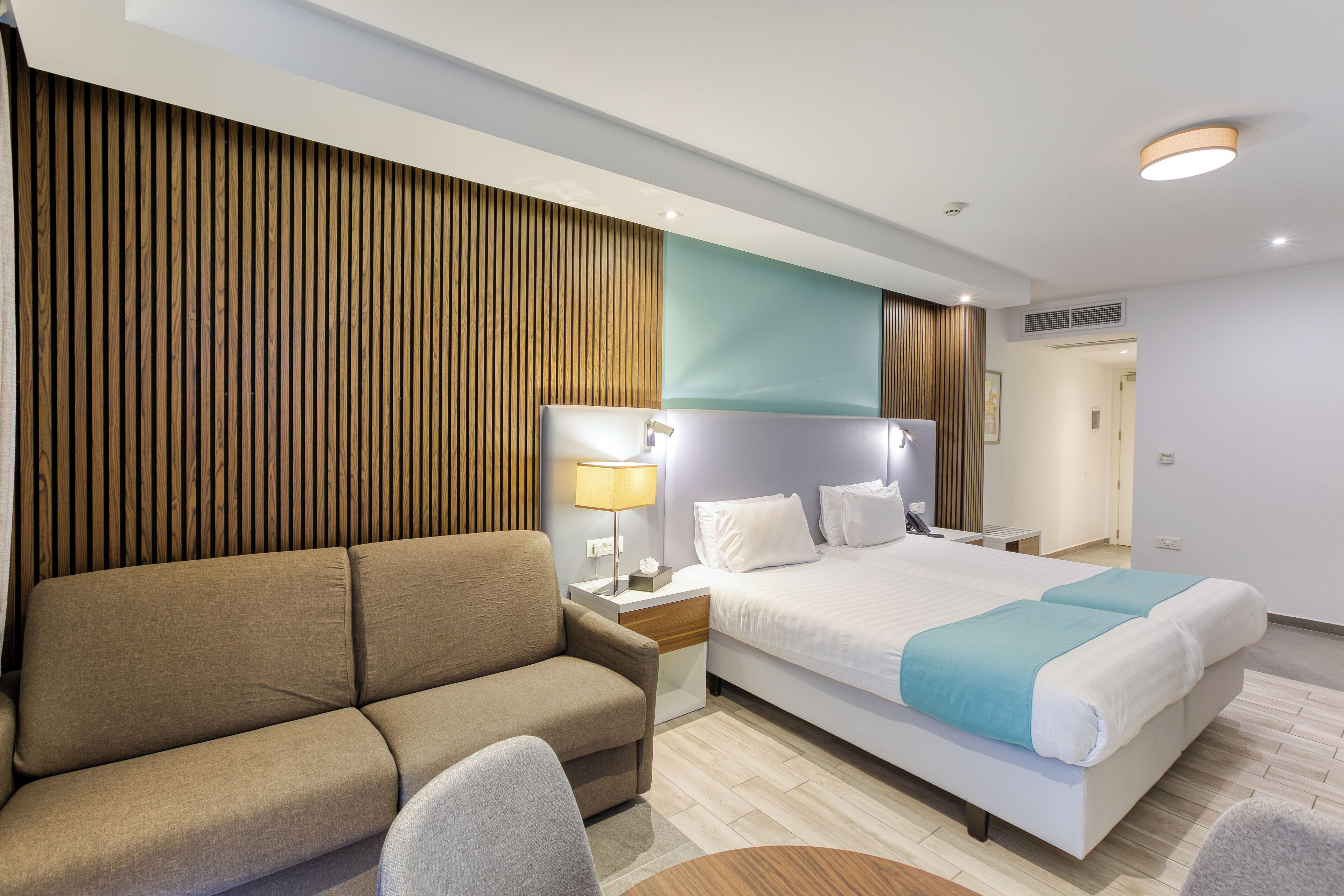 A modern hotel room with a large bed, a sofa, a desk, and a lamp.