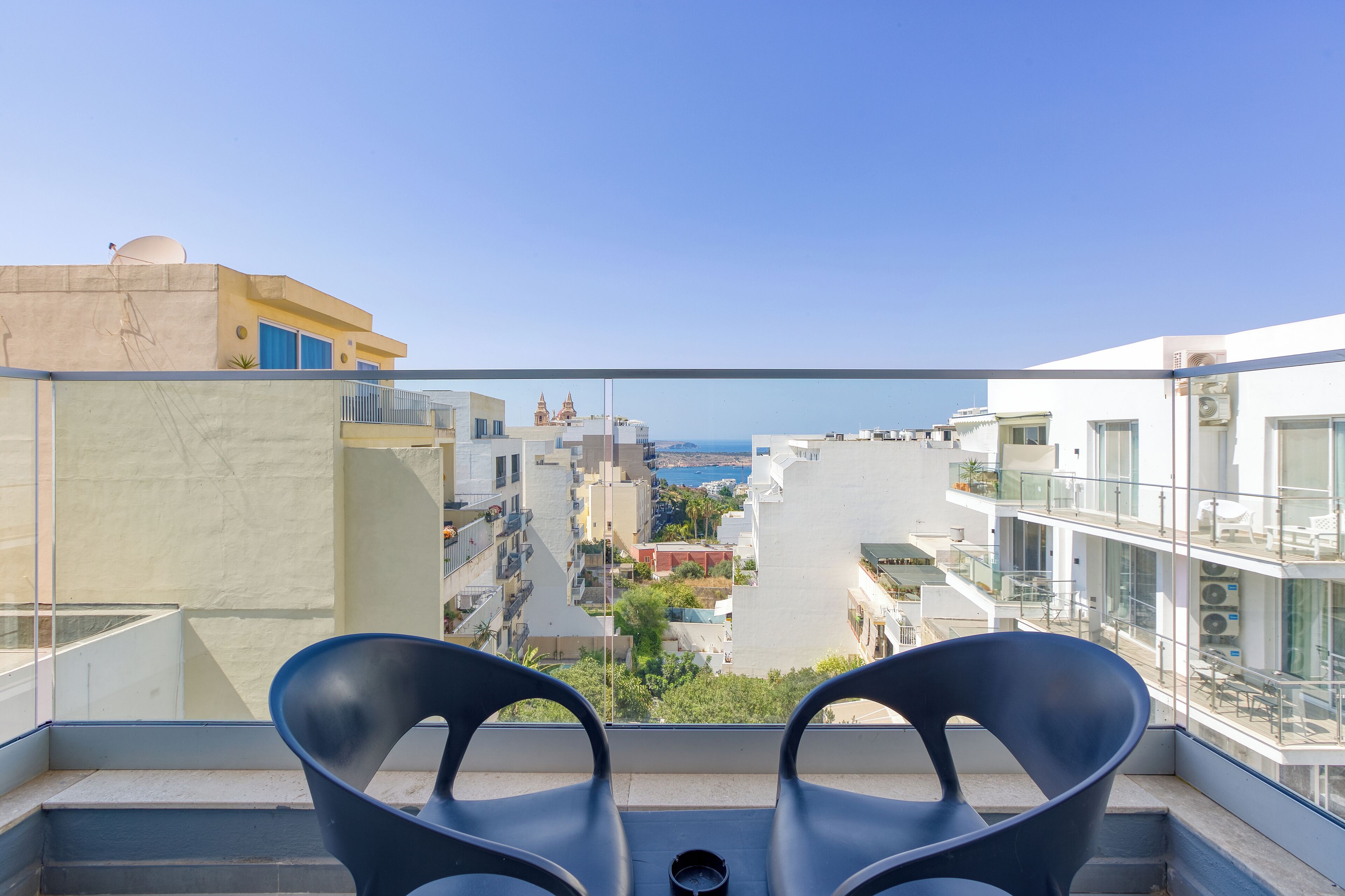 A balcony with two chairs, a clear view of the sea, and a cityscape in the background.