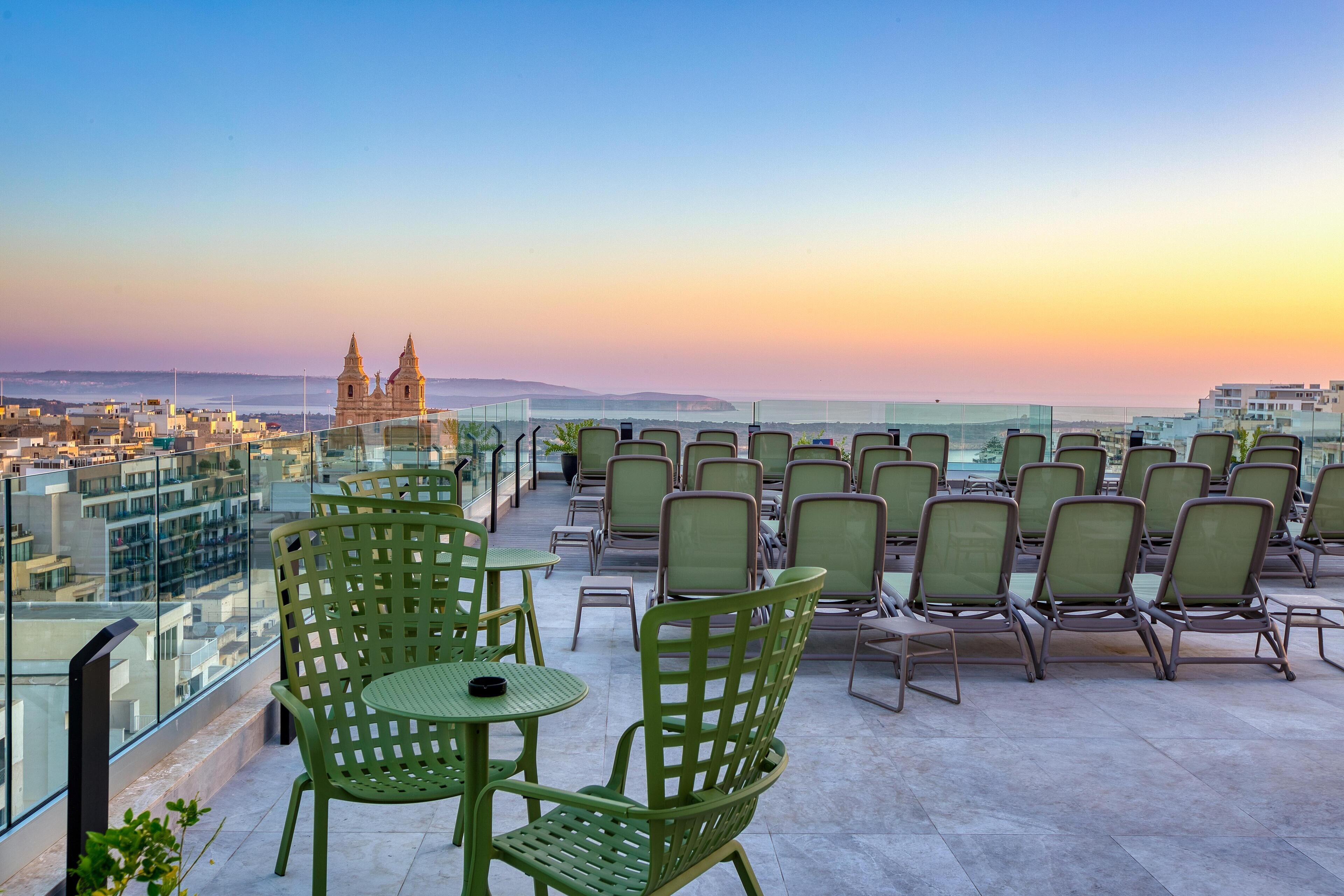 A rooftop terrace with green chairs and tables, offering a view of a coastal cityscape and a prominent church.