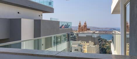 Um edifício moderno com varanda de vidro que oferece vista para uma paisagem costeira, incluindo uma catedral imponente.
