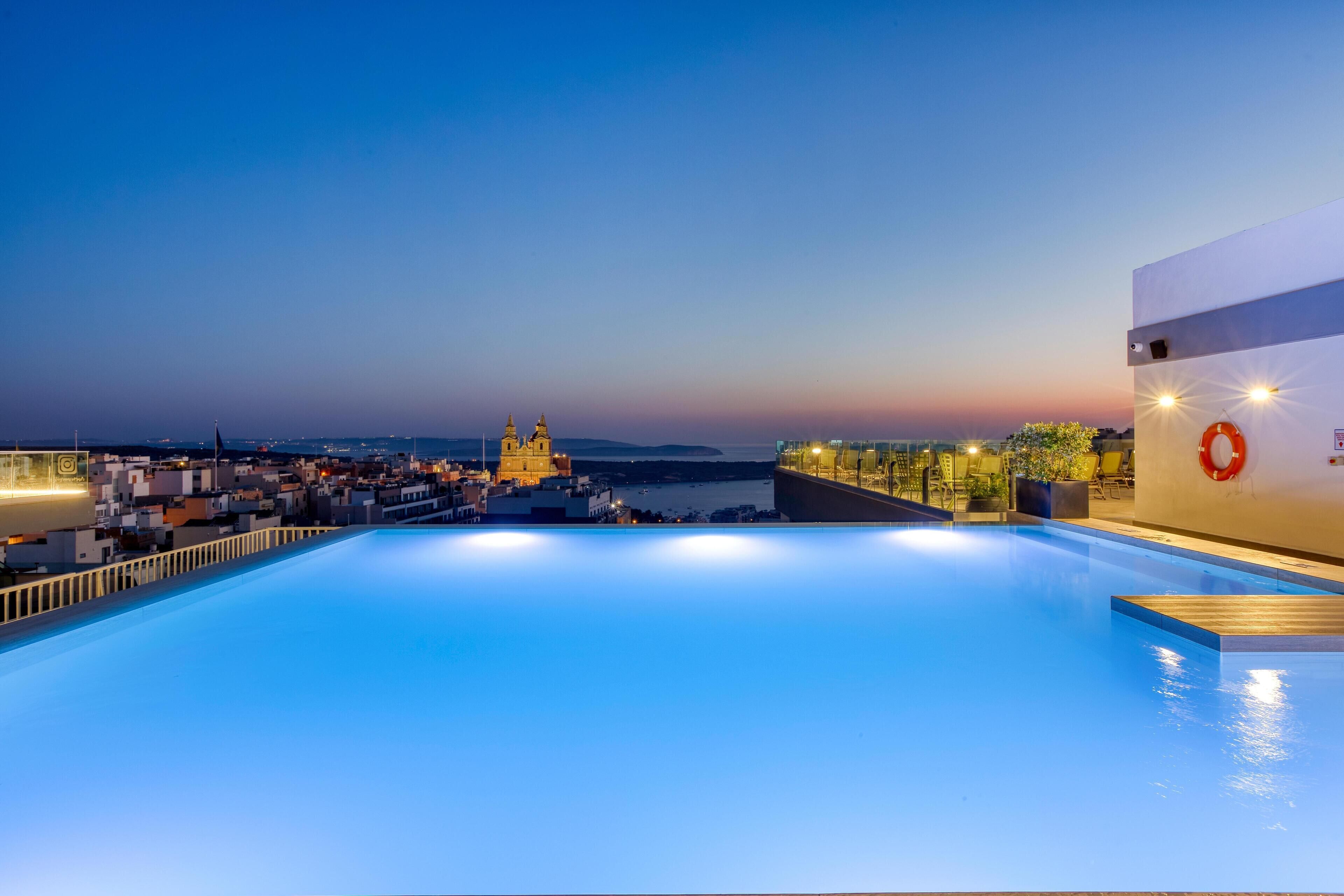 A rooftop pool with a city view, a lifebuoy on the wall, and a sunset in the background.