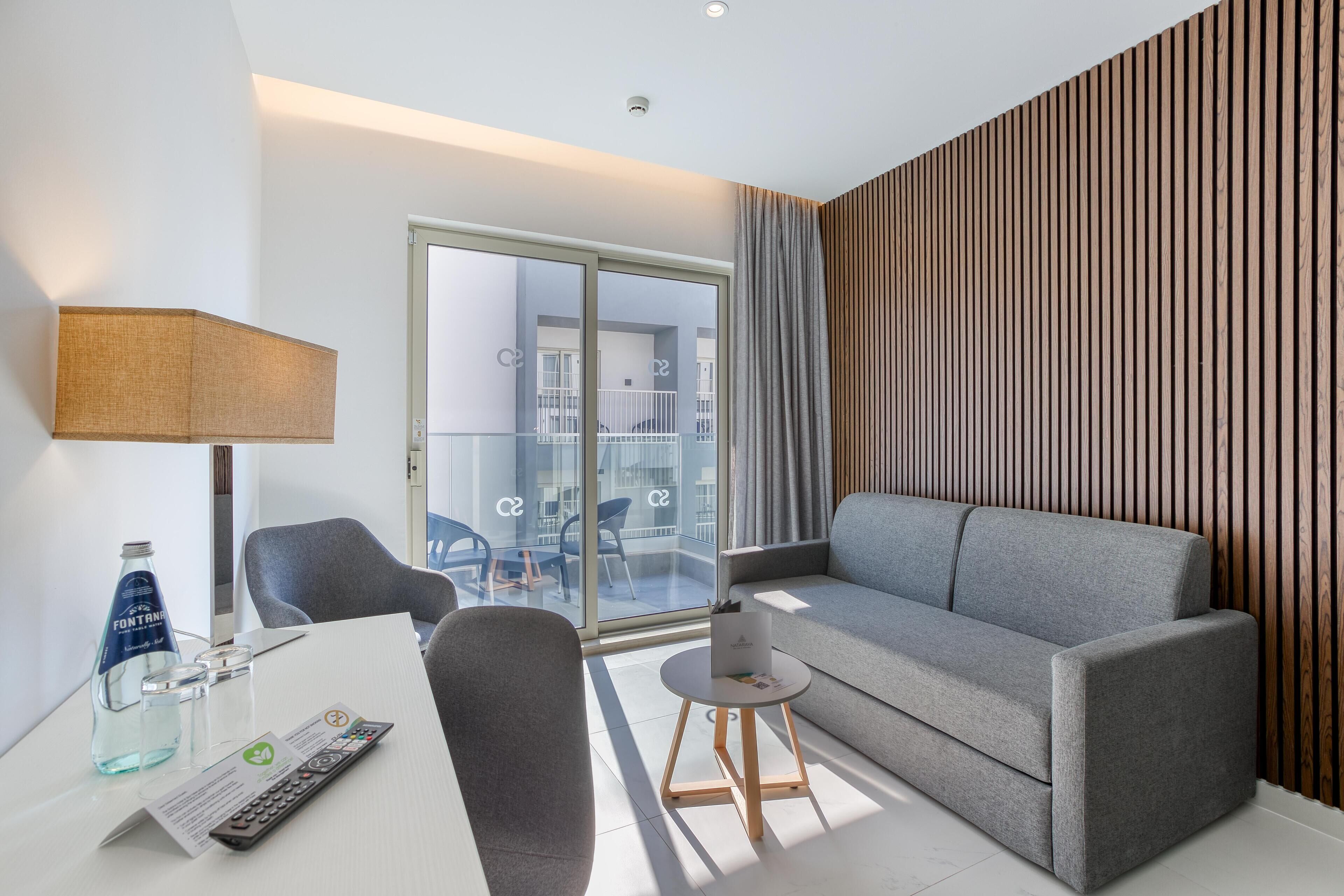 Moderni hotellihuone, jossa on harmaa sohva, pieni pyöreä pöytä, lamppu ja lasiovi, joka johtaa parvekkeelle.