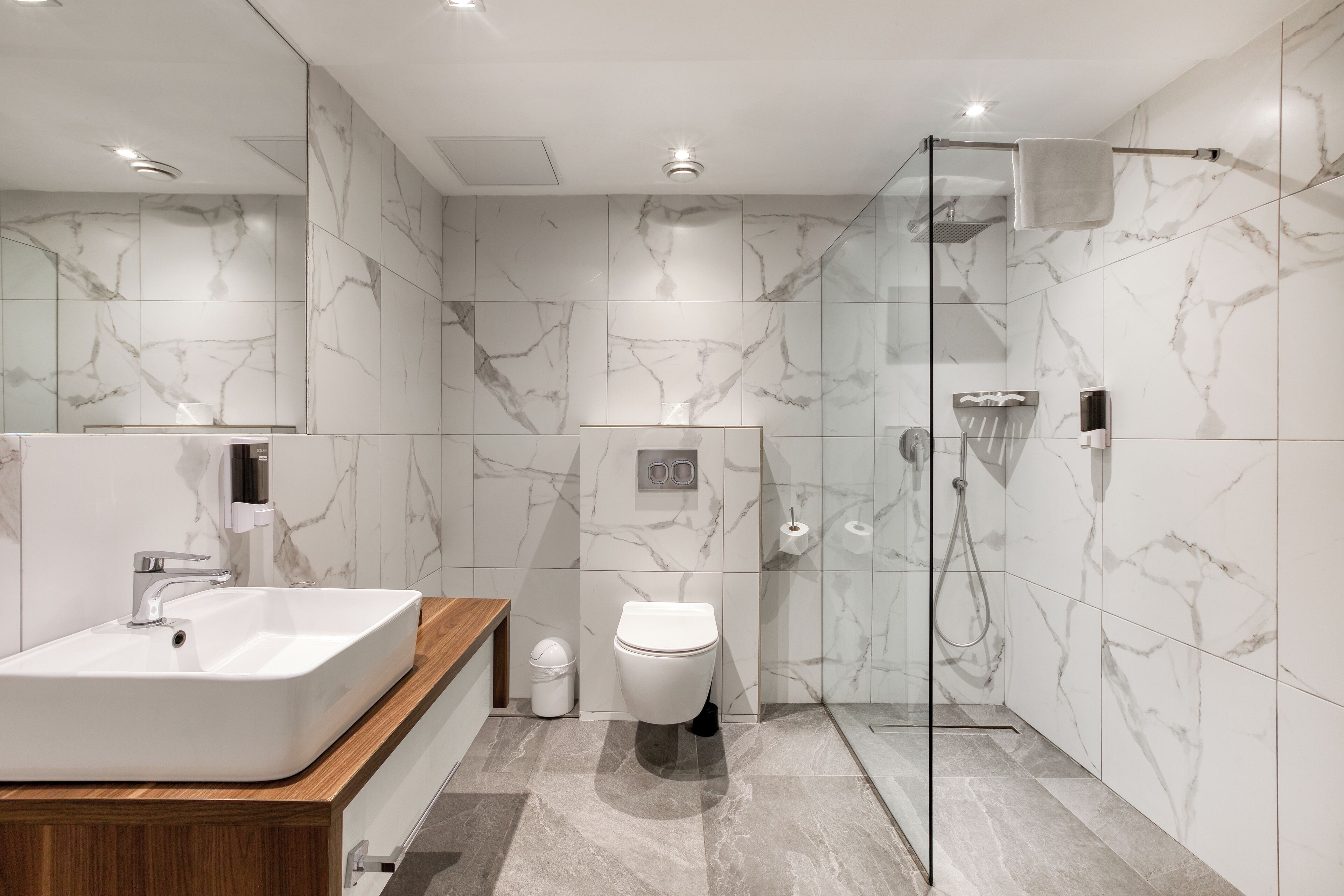 A modern bathroom with a marble wall, a wooden vanity, a white sink, a toilet, and a glass-enclosed shower area.