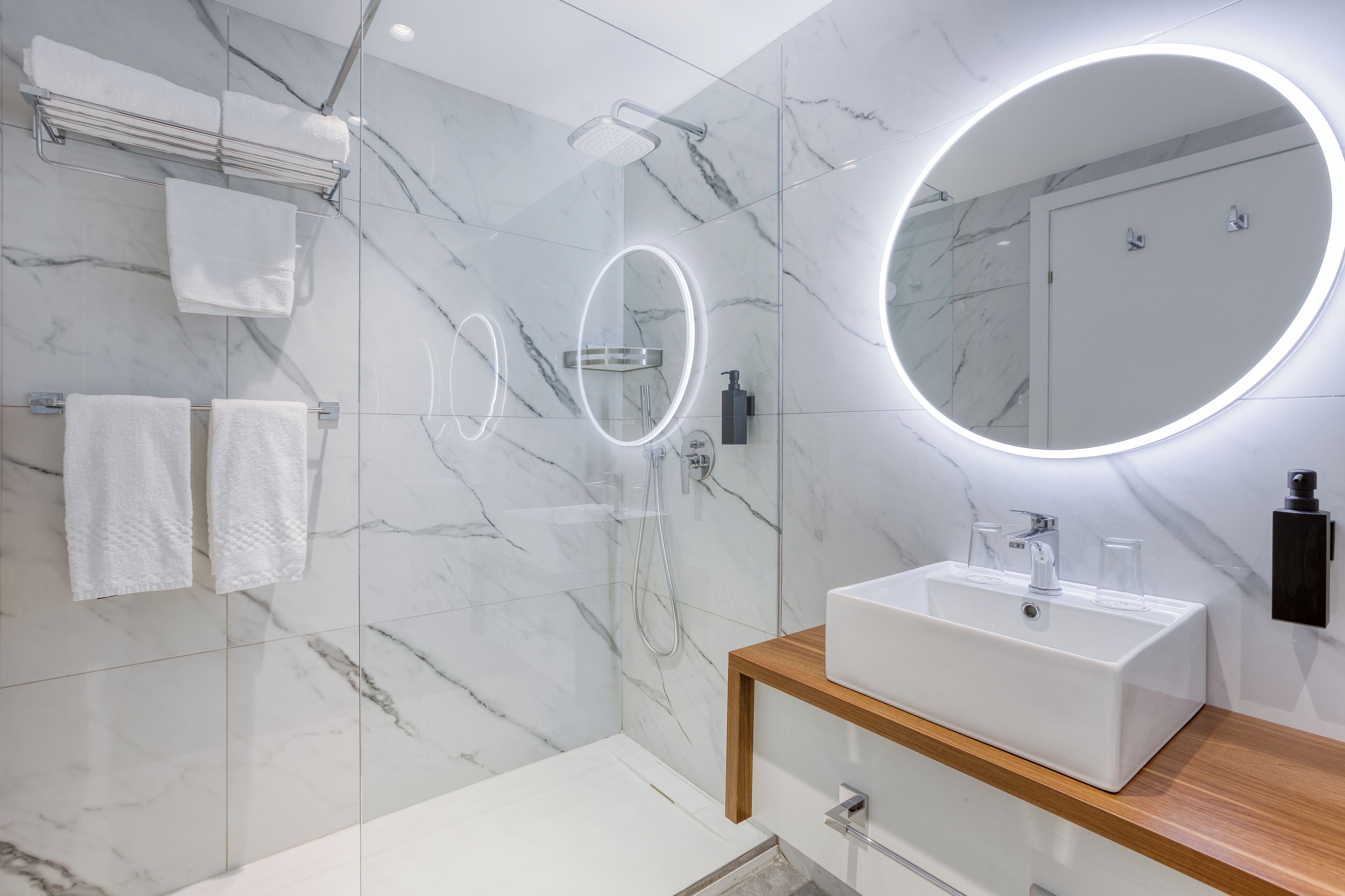 A modern bathroom with a marble wall, a round mirror, a white sink, and a shower.