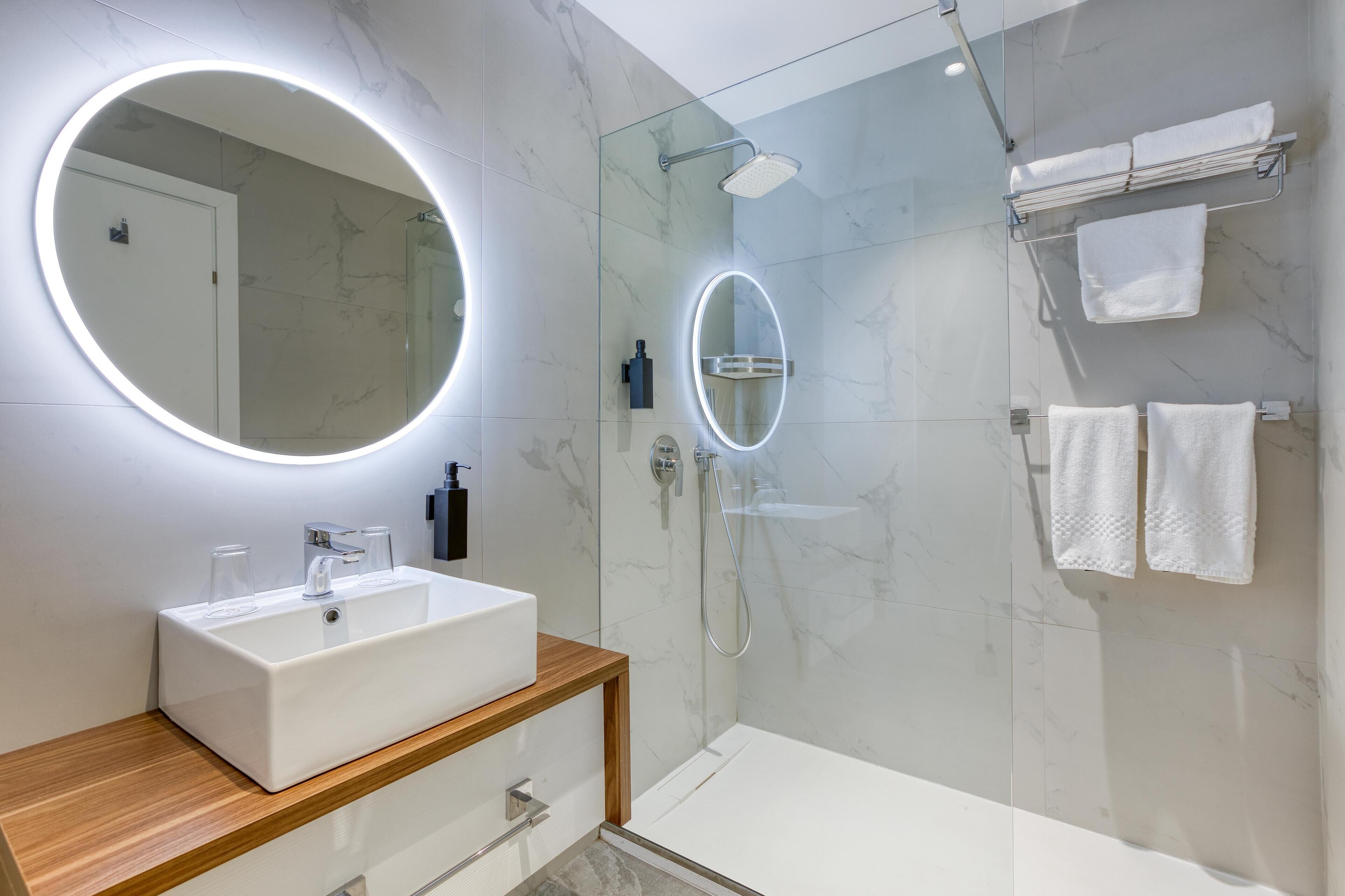 Ein modernes Badezimmer mit einem weißen Waschbecken, zwei Spiegeln, einer Duschkabine und weißen Handtüchern an einem Handtuchhalter.