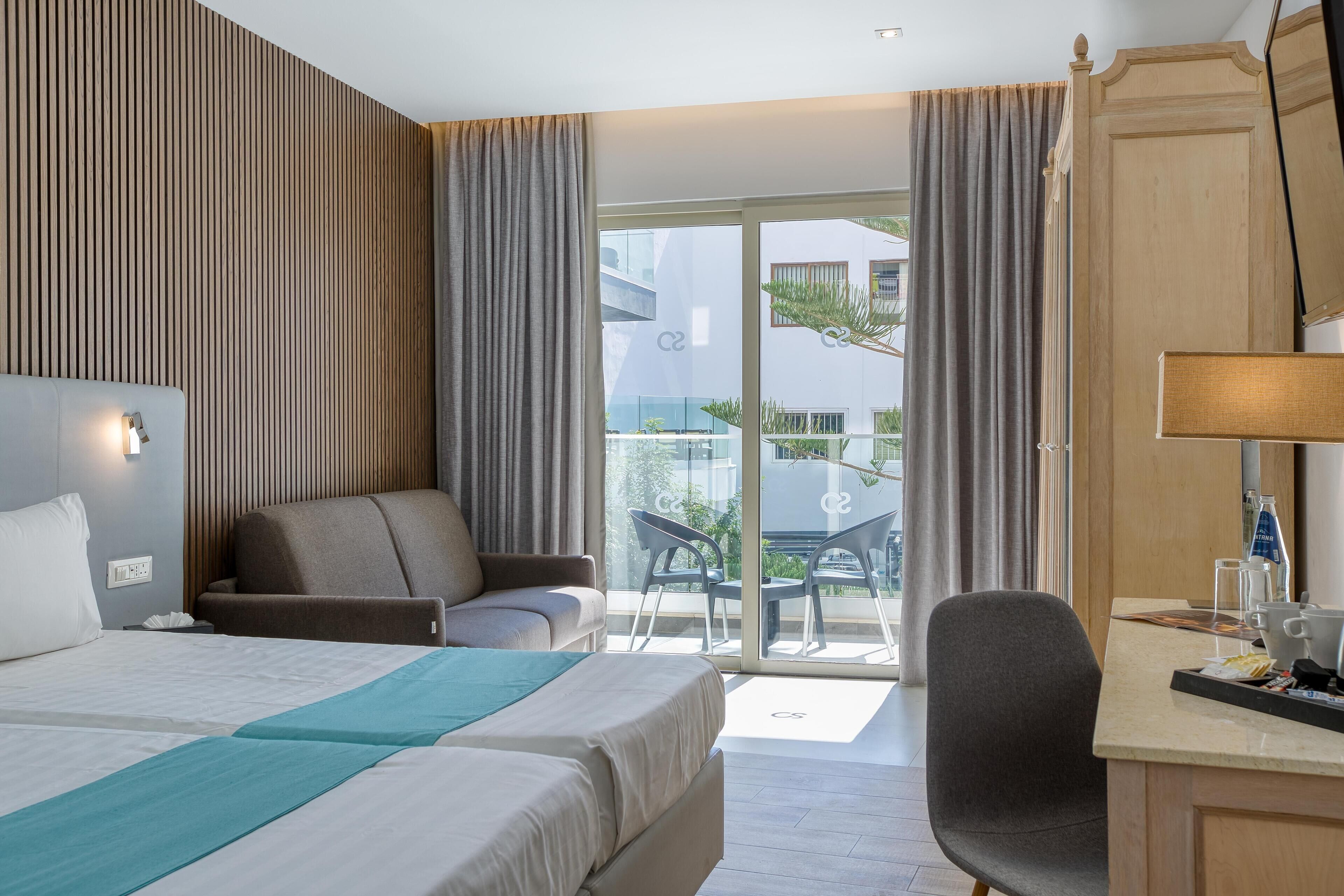 Habitación de hotel con cama, escritorio, silla y balcón con vistas a edificios.