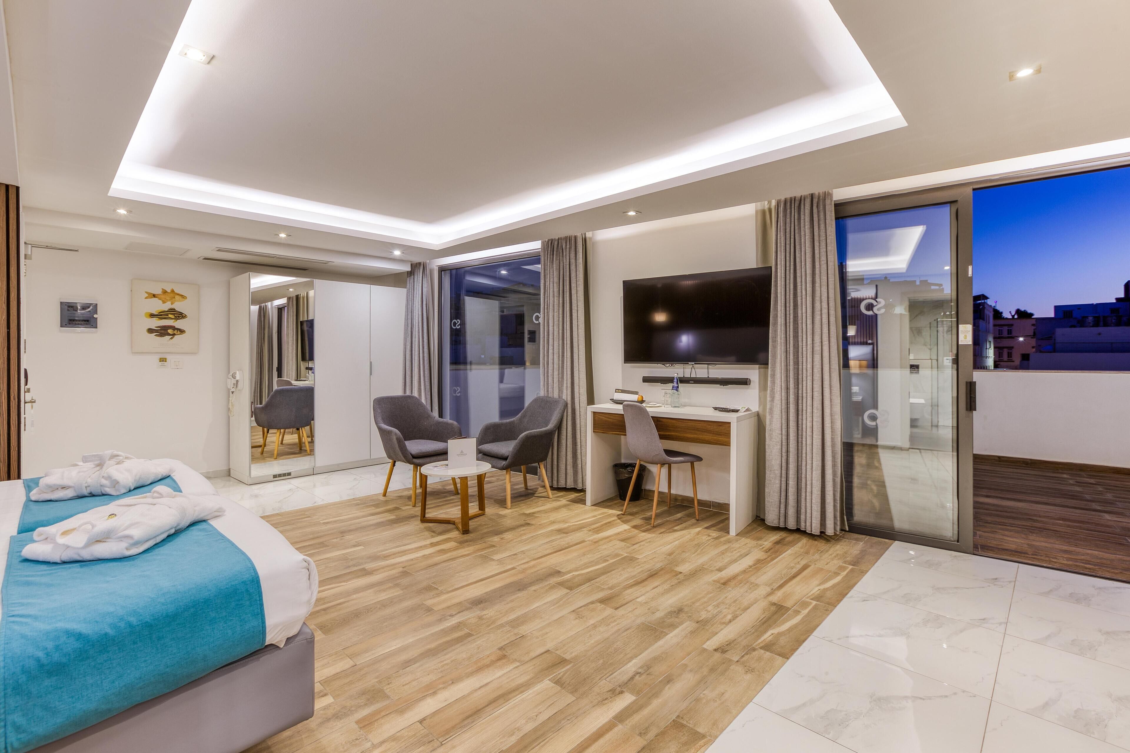 Moderni hotellihuone, jossa on sänky, työpöytä, tuoleja ja televisio.