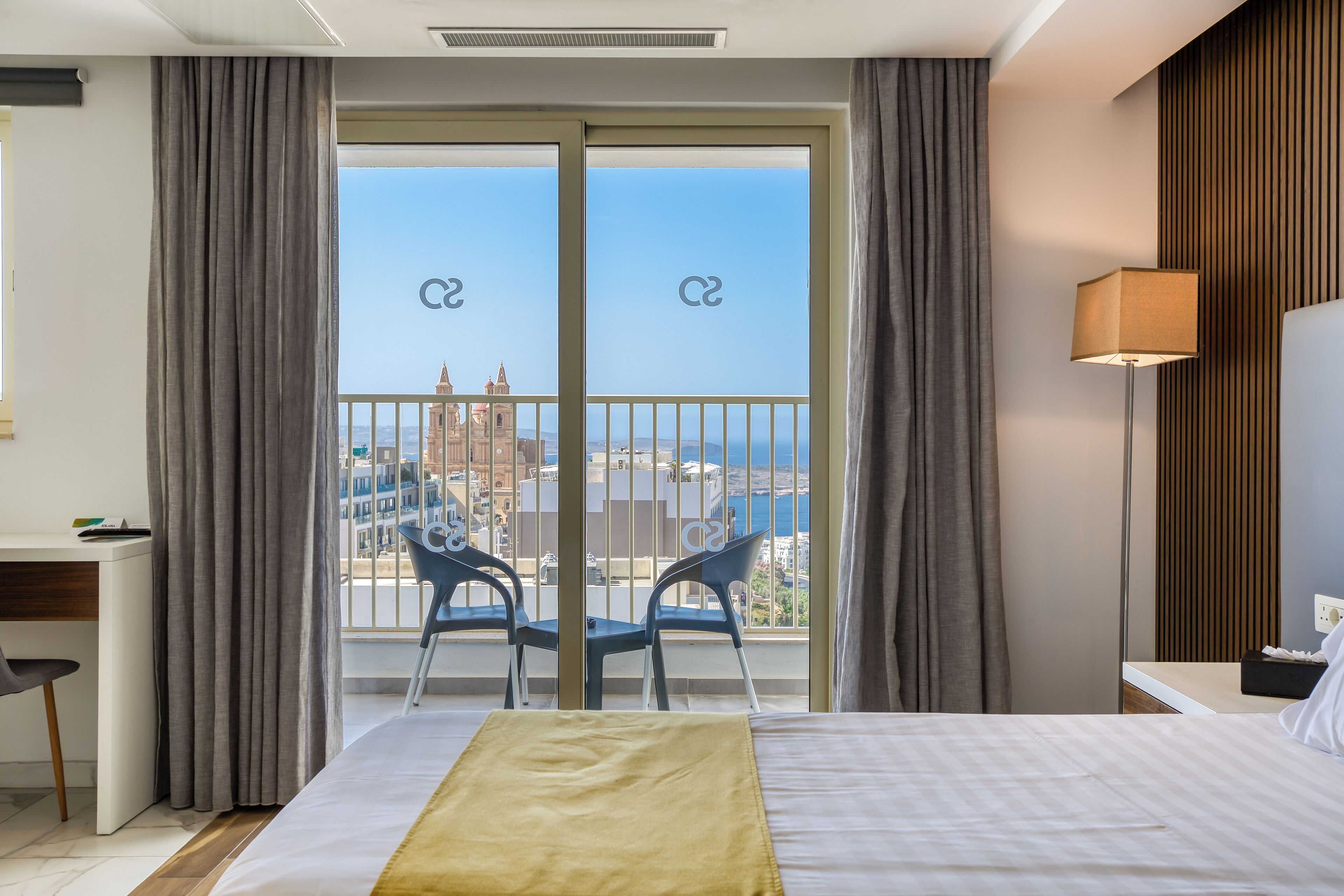 Quarto de hotel com uma cama, uma escrivaninha, uma cadeira e vista para a cidade e para o mar através de grandes janelas.