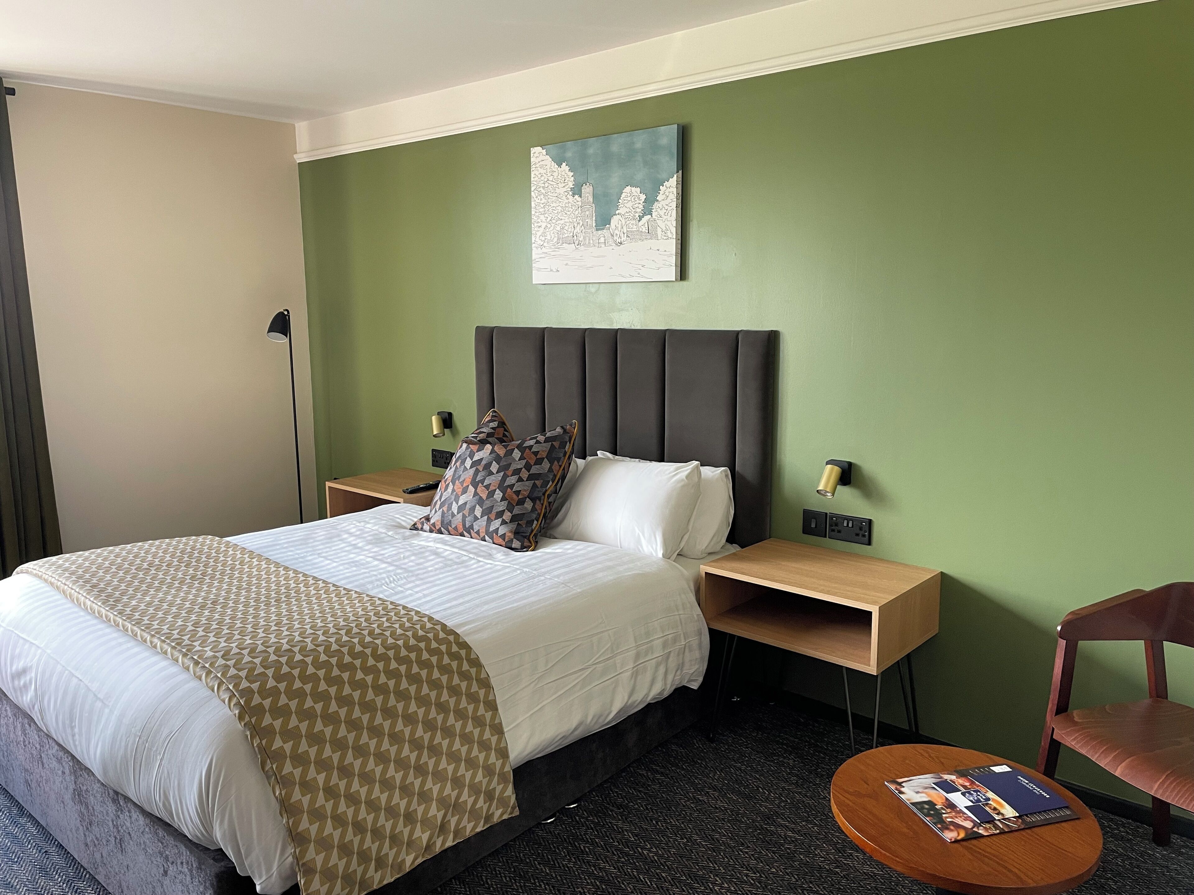 A hotel room with a bed, a nightstand, a lamp, a chair, and a small table with a magazine.