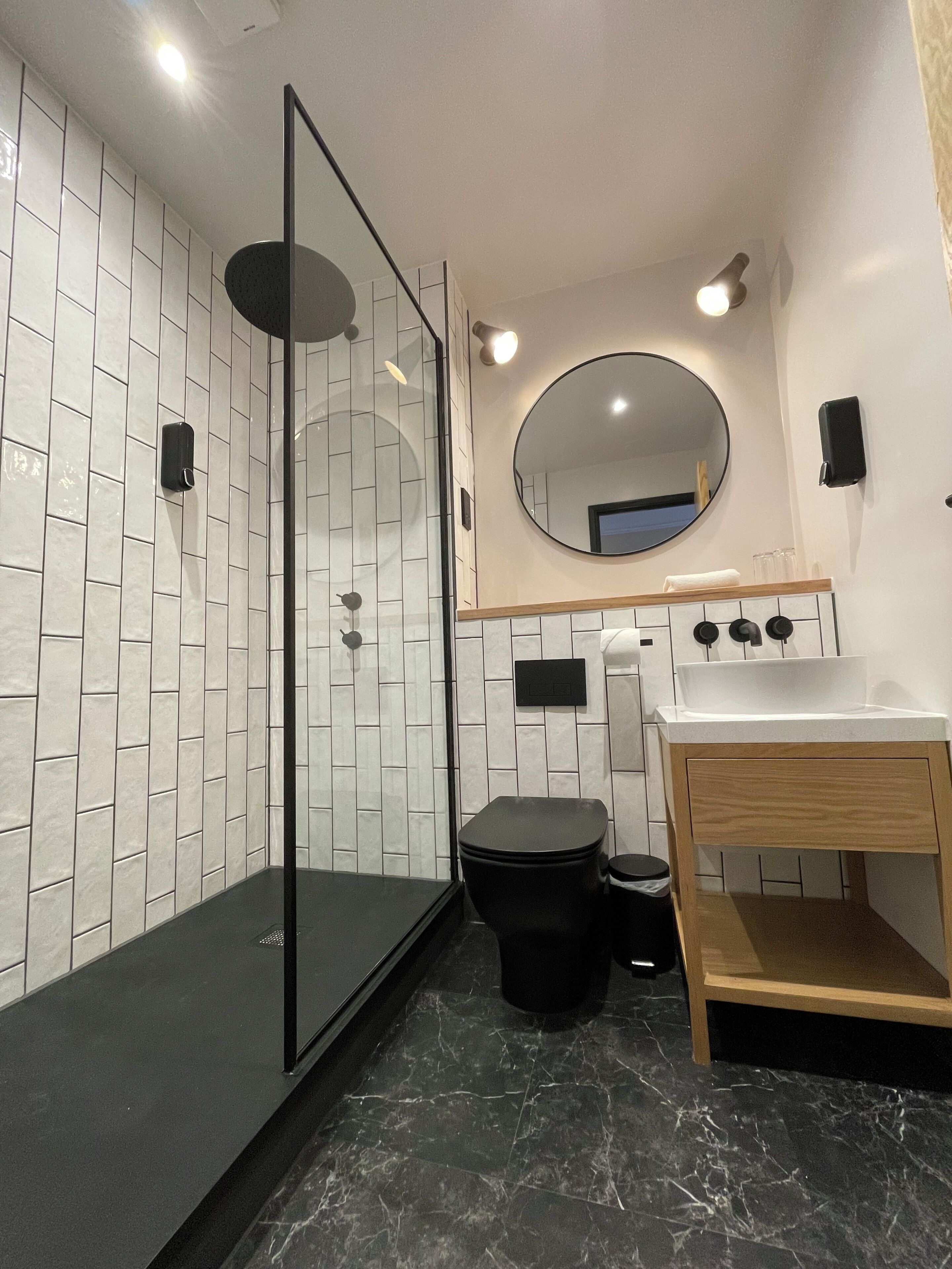 Un baño moderno con cabina de ducha de vidrio, un inodoro, un lavabo con tocador de madera y un espejo redondo sobre el lavabo.