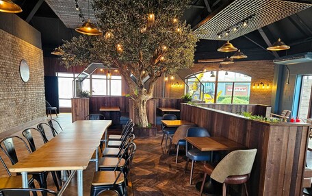 A restaurant interior with wooden tables and chairs, a large tree centerpiece, and a brick wall.