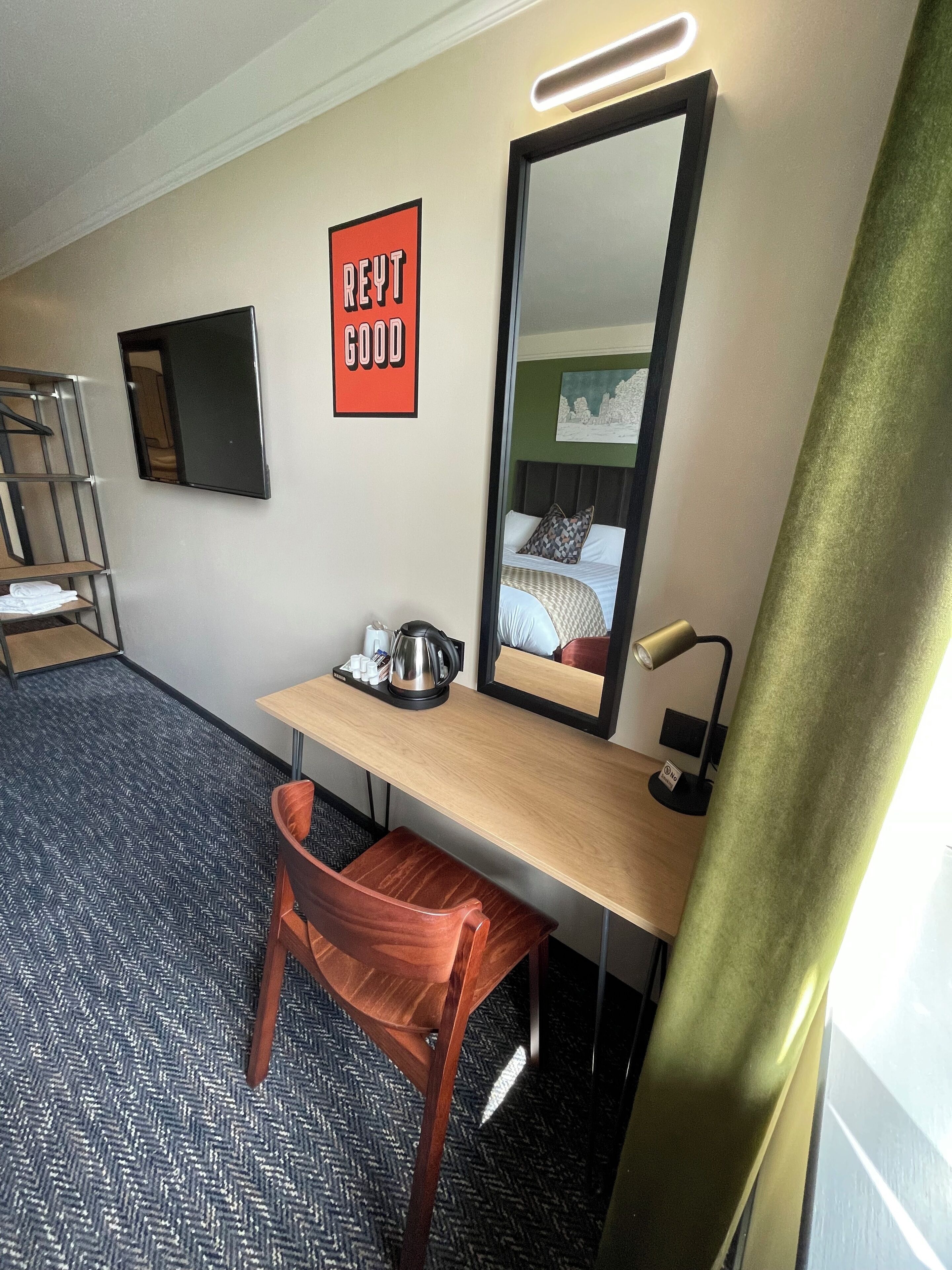 Ett hotellrum med en säng, ett skrivbord med en vattenkokare, en stol, en spegel och en skylt på väggen med texten ”REYT GOOD”.