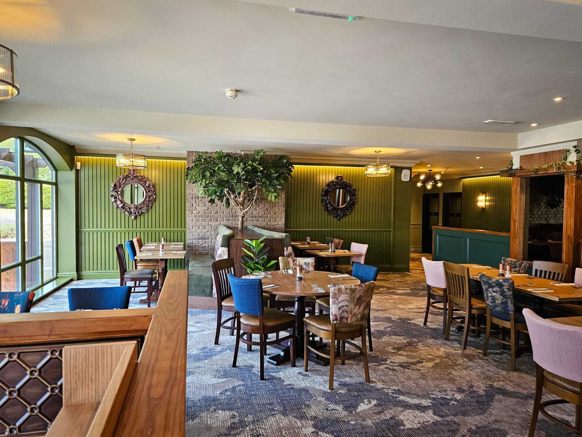 A restaurant with wooden tables and chairs, a large mirror, and a green wall with a plant.