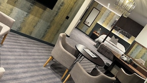 Una habitación de hotel moderna con un televisor de pantalla plana grande montado en la pared, una zona de estar cómoda con sofás y sillones, una mesa de comedor y una araña.
