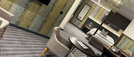 A modern hotel room with a large flat-screen TV mounted on the wall, a comfortable seating area with sofas and chairs, a dining table, and a chandelier.