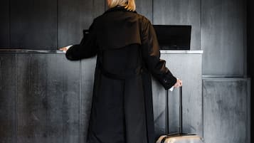 A person in a black coat standing at a reception desk with a suitcase.