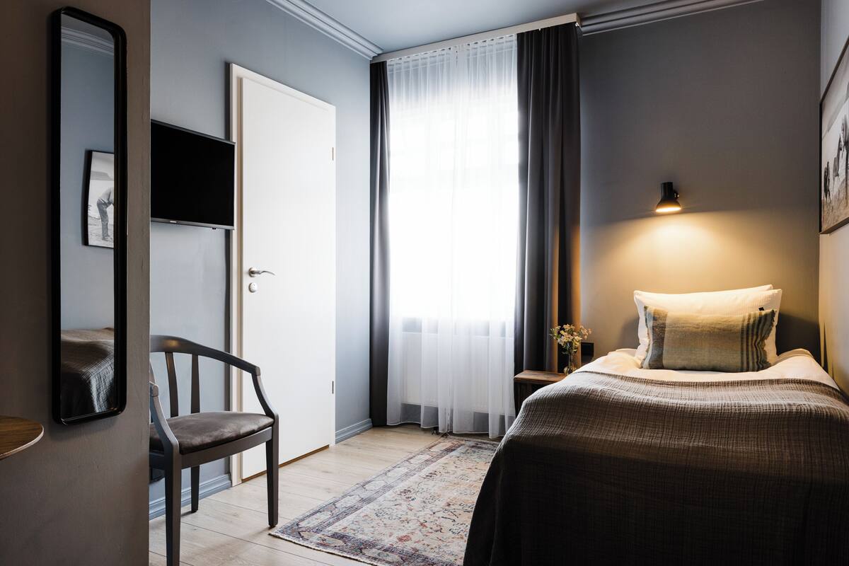 Quarto de hotel com cama, cadeira, televisão e janela com cortinas.