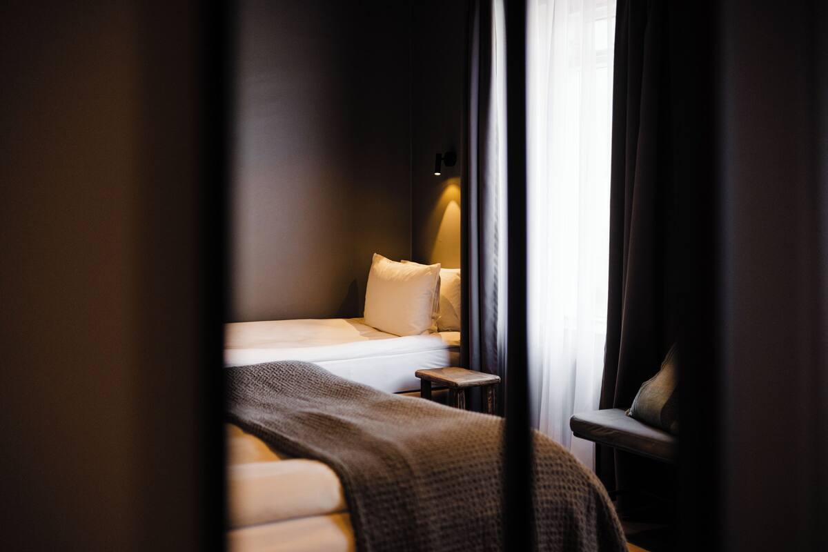 Quarto de hotel com cama, mesinha de cabeceira, cadeira e janela com cortinas.