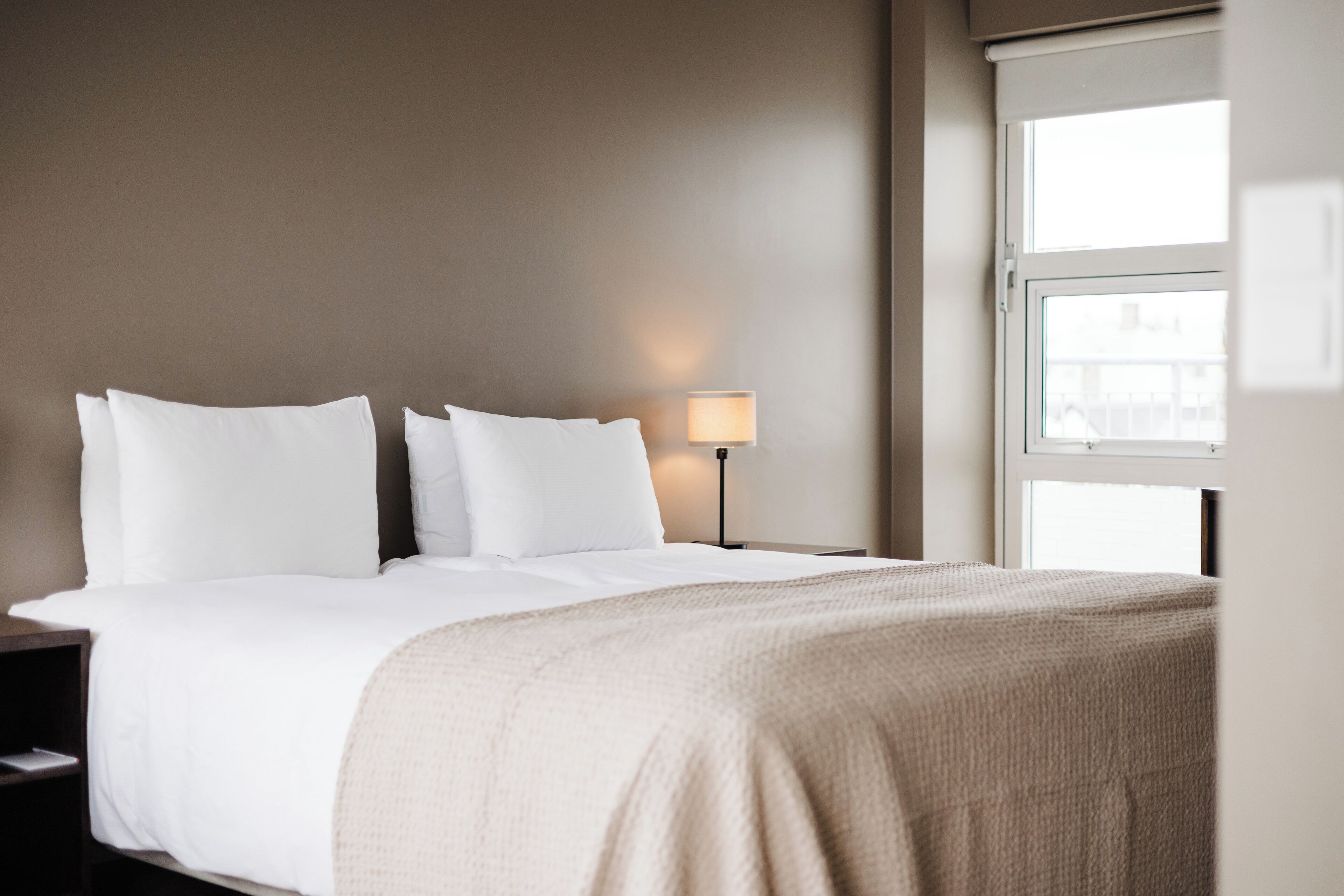 A neatly made bed with white bedding and a beige throw blanket, a bedside lamp, and a window with a view of buildings.