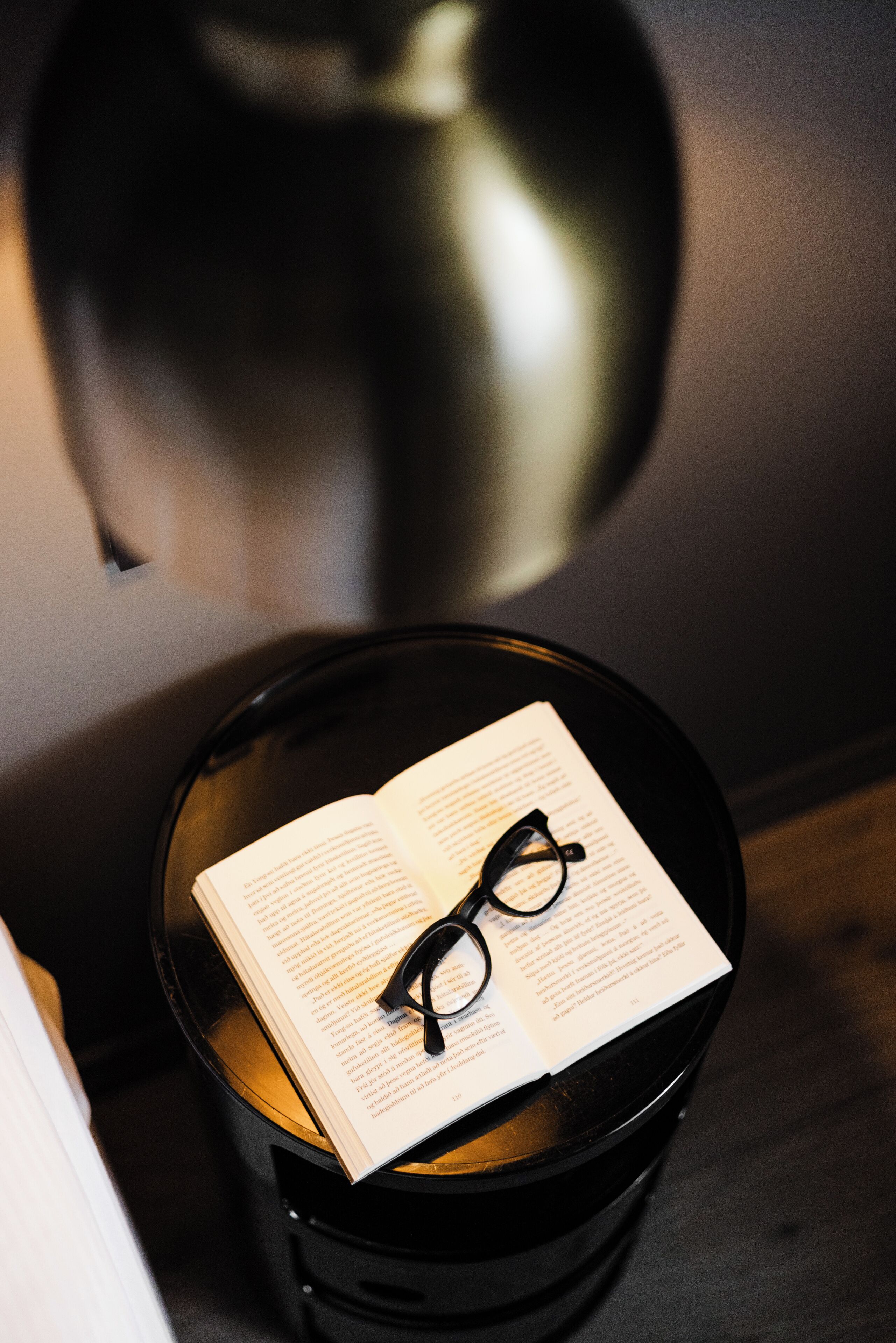 Ein Buch und eine Brille auf einem schwarzen Tisch, im Hintergrund steht eine Lampe.