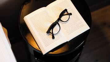 Ein Buch und eine Brille auf einem schwarzen Tisch, im Hintergrund steht eine Lampe.