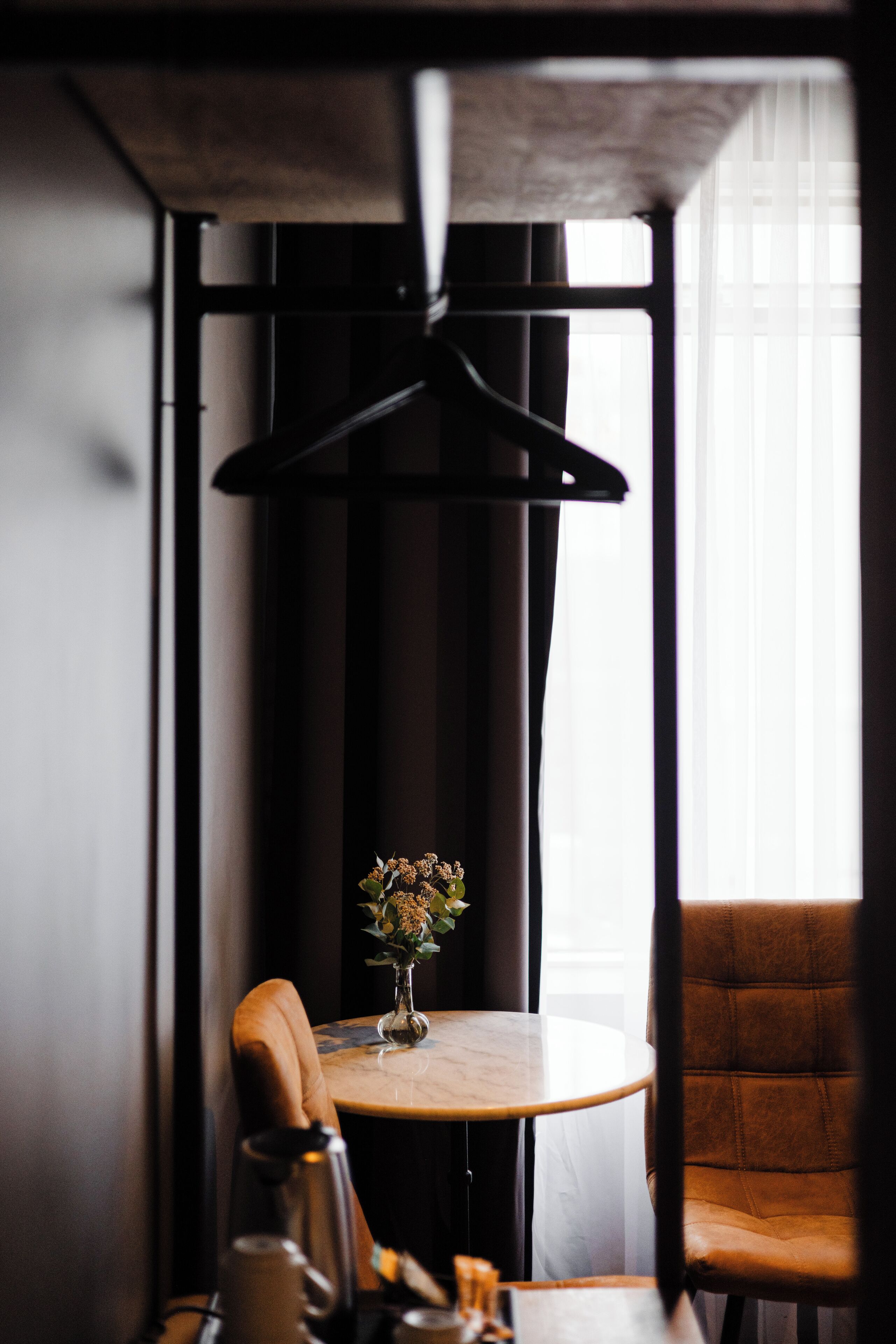 Una pequeña mesa redonda con un florero, un sillón de cuero marrón y una lámpara colgante en una habitación con ventana y cortinas.