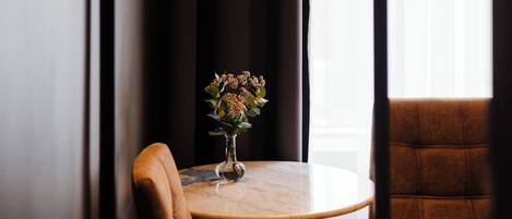 Una pequeña mesa redonda con un florero, un sillón de cuero marrón y una lámpara colgante en una habitación con ventana y cortinas.