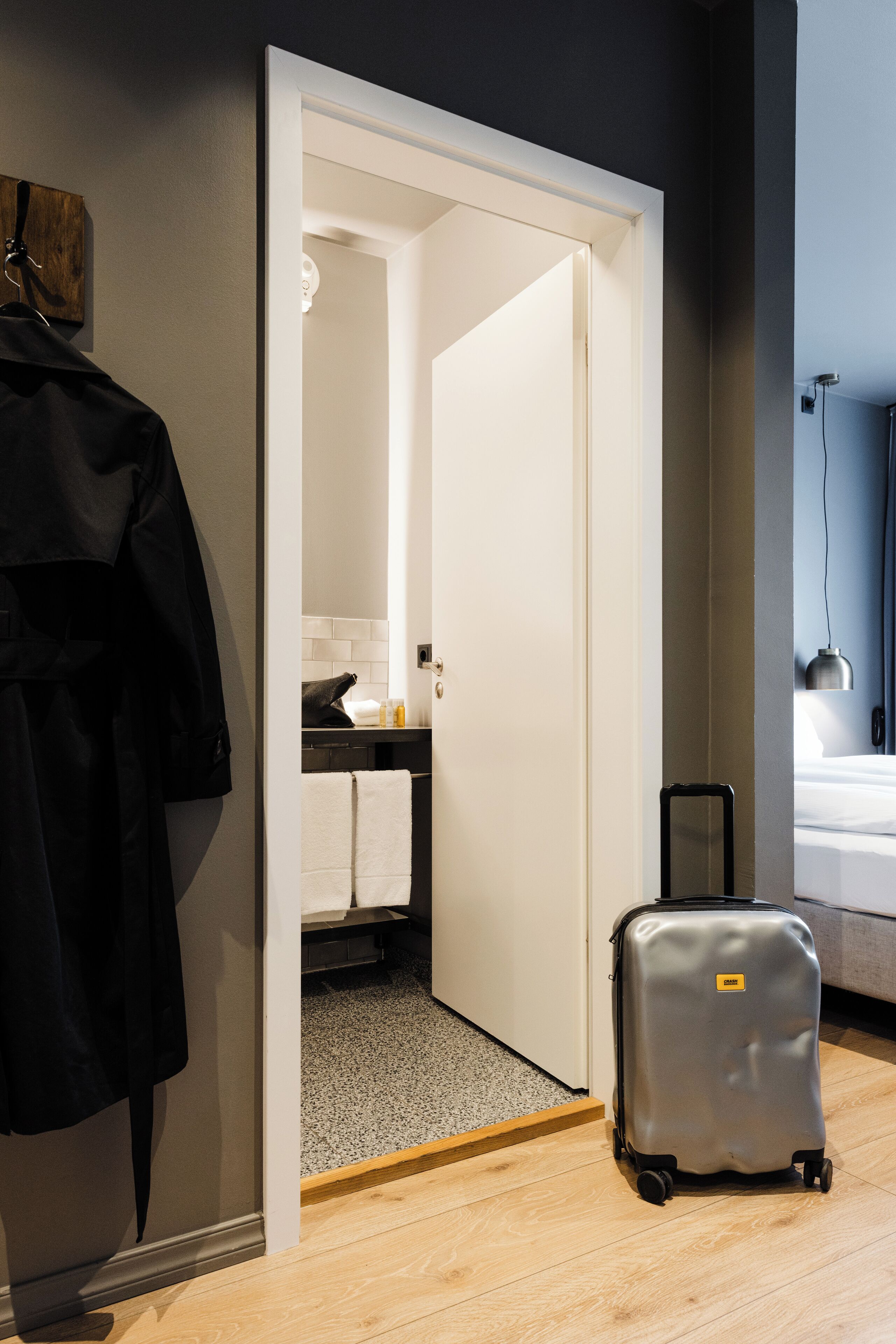 Una habitación de hotel con piso de madera, un abrigo negro colgado en la pared, una maleta en el suelo y una cama con cabecera blanca.