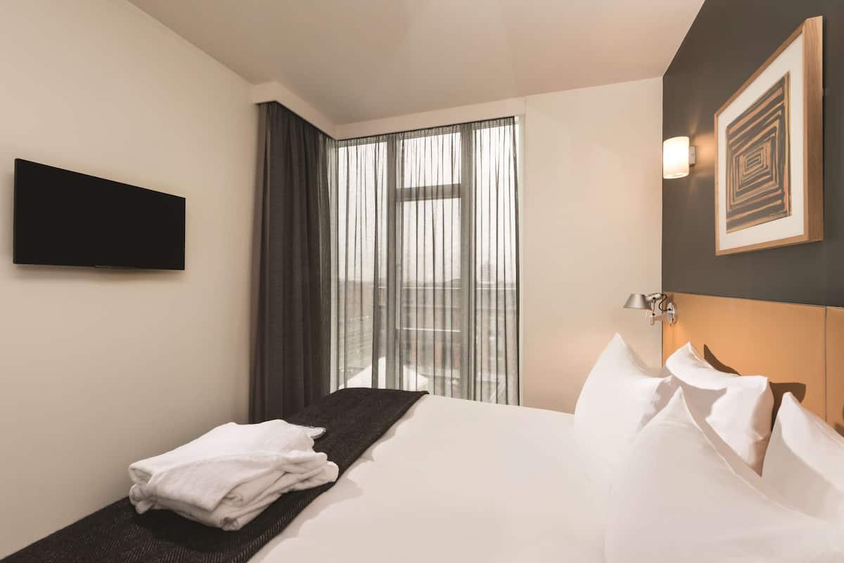 Ein Hotelzimmer mit einem Bett, einem an der Wand befestigten Flachbildfernseher, einem Fenster mit durchscheinenden Vorhängen und einem gerahmten Kunstwerk an der Wand.