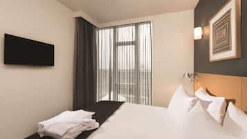Ein Hotelzimmer mit einem Bett, einem an der Wand befestigten Flachbildfernseher, einem Fenster mit durchscheinenden Vorhängen und einem gerahmten Kunstwerk an der Wand.