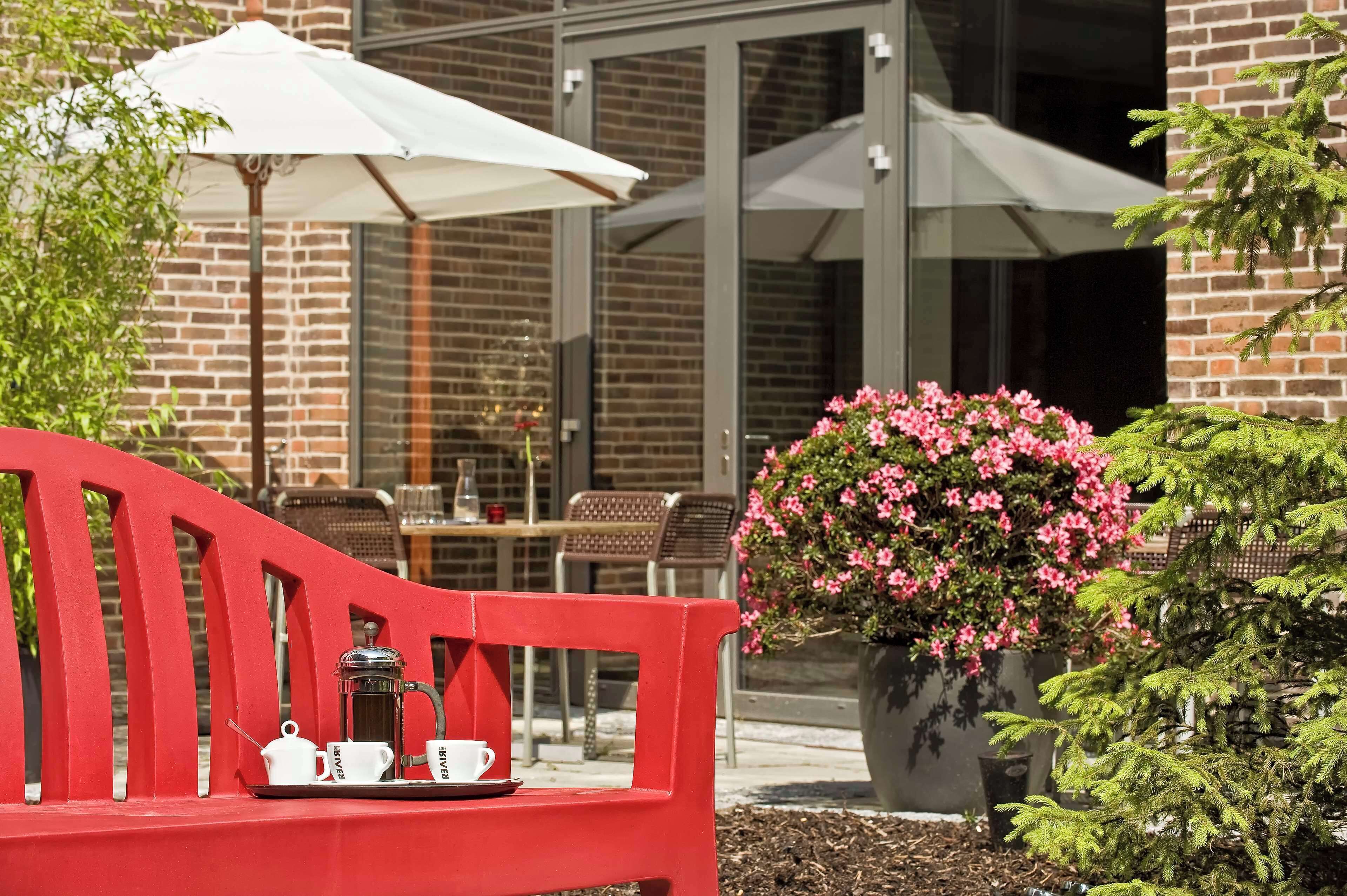 A red bench with a tray of coffee and cups, a white umbrella, and a potted plant with pink flowers.
