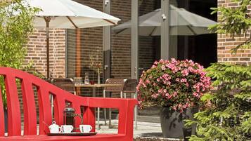 A red bench with a tray of coffee and cups, a white umbrella, and a potted plant with pink flowers.