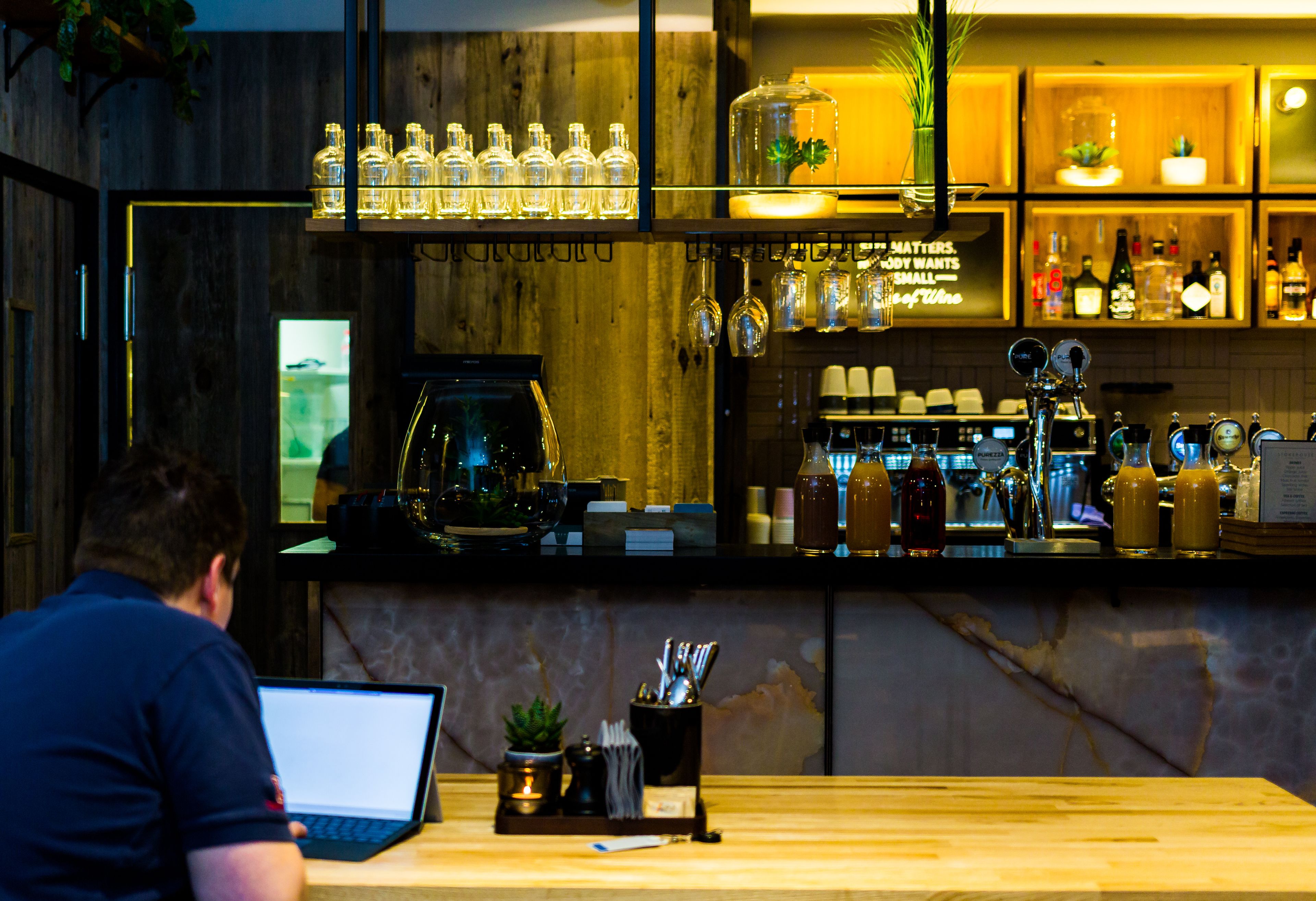 Una zona de bar con una computadora portátil sobre una barra, estantes con botellas y una máquina de café.