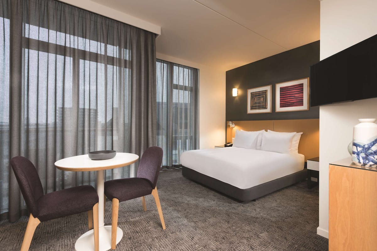 Ein Hotelzimmer mit einem Bett, zwei Stühlen, einem kleinen runden Tisch, einem Nachttisch und einem an der Wand montierten Fernseher.