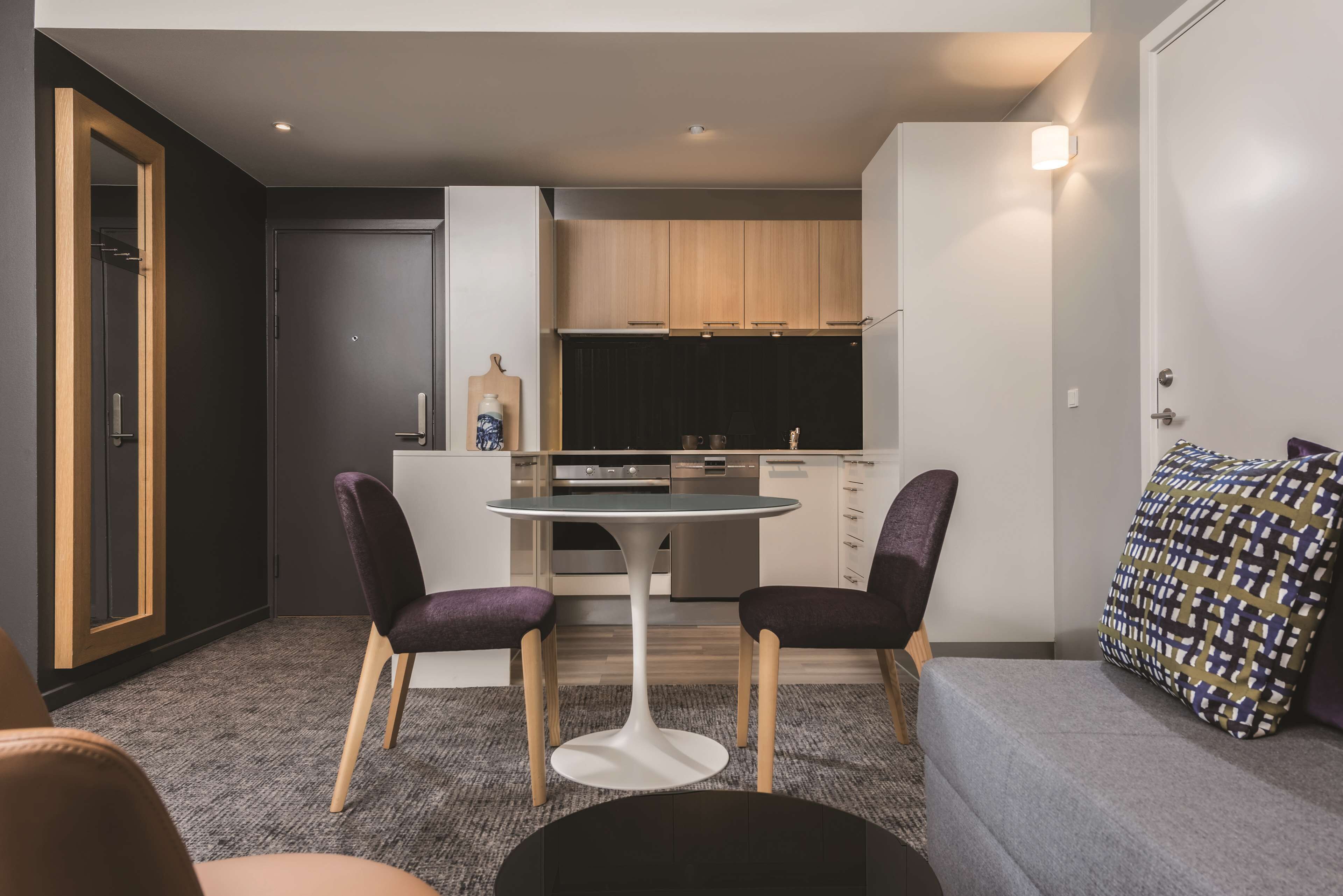 Un espacio habitable compacto con kitchenette, dos sillas moradas, una mesa redonda y un sofá gris.