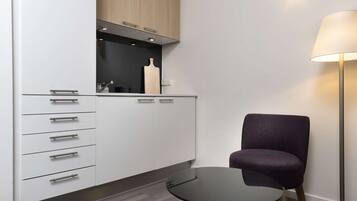 Une cuisine moderne avec des meubles blancs, un plan de travail noir et une planche à découper en bois. On y voit une table ronde avec une chaise, et une lampe sur pied est visible.