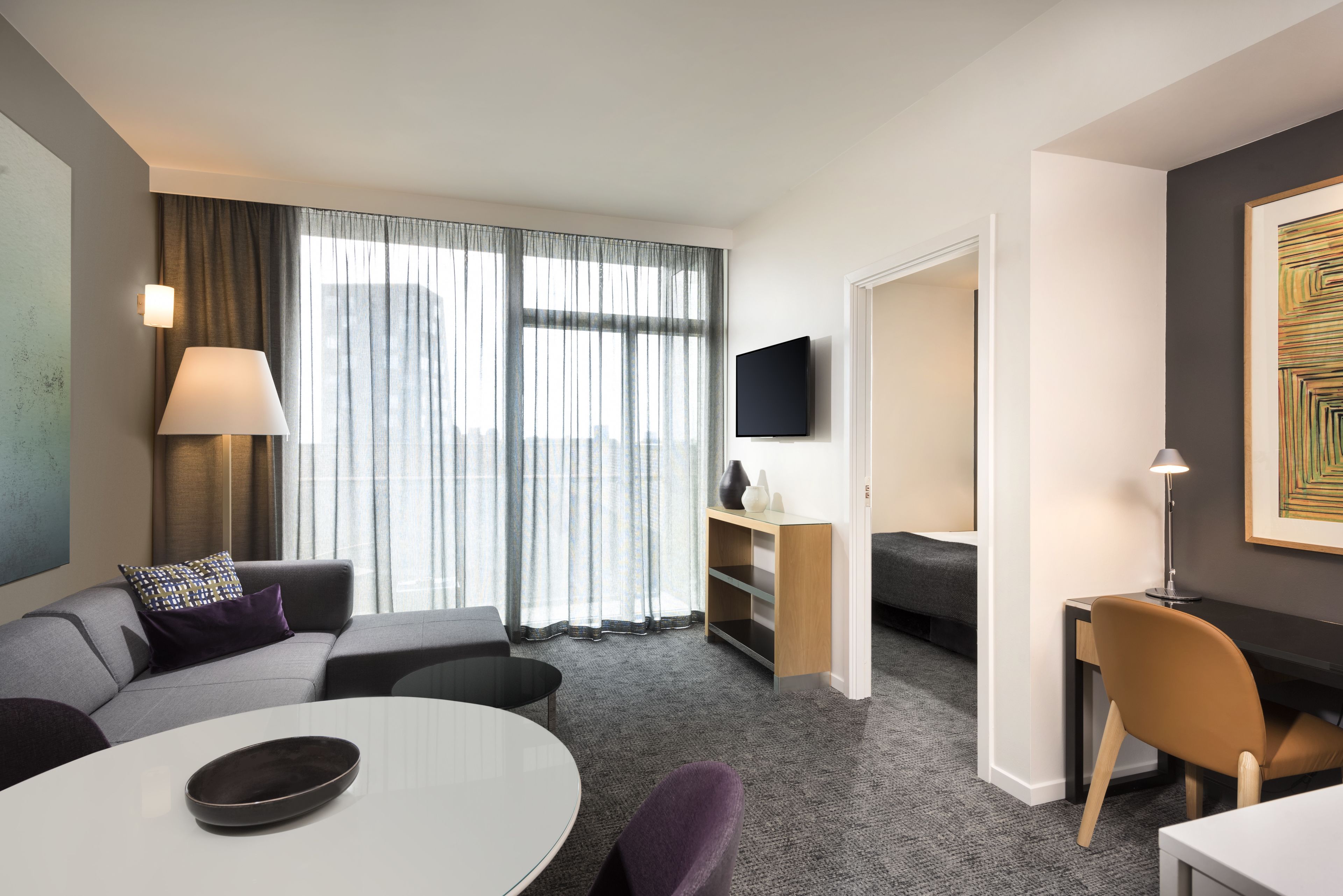 Une chambre d’hôtel moderne avec un canapé gris, une table de salle à manger ronde, un téléviseur à écran plat fixé au mur et une grande fenêtre ornée de rideaux légers.