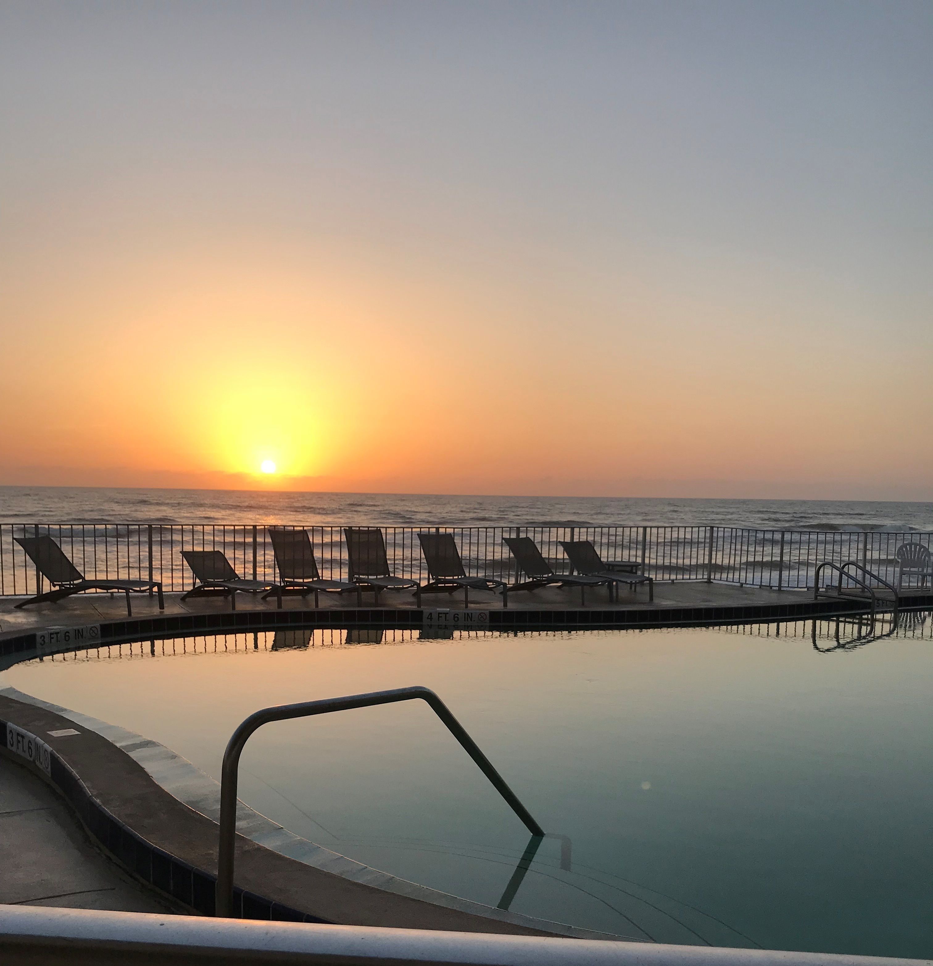 A poolside area with lounge chairs and a view of the sunset over the ocean.