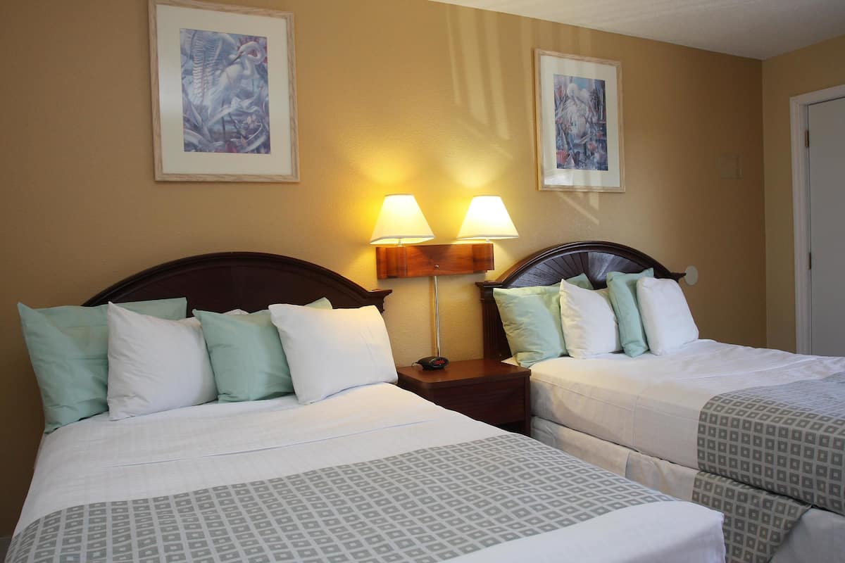 A hotel room with two beds, each with a wooden headboard, white bedding, and a patterned bedspread. There are two wall-mounted lamps on a wooden nightstand between the beds.