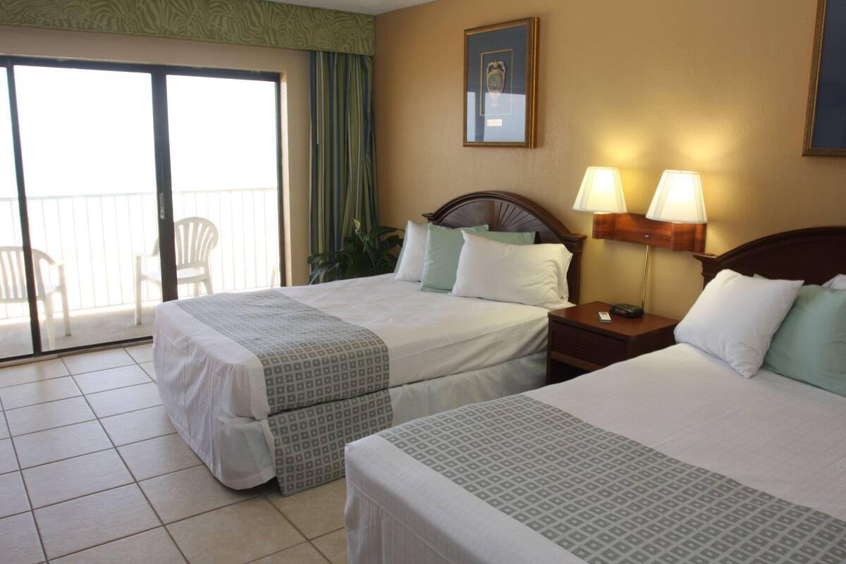 A hotel room with two beds, a balcony with chairs, and a nightstand with lamps.