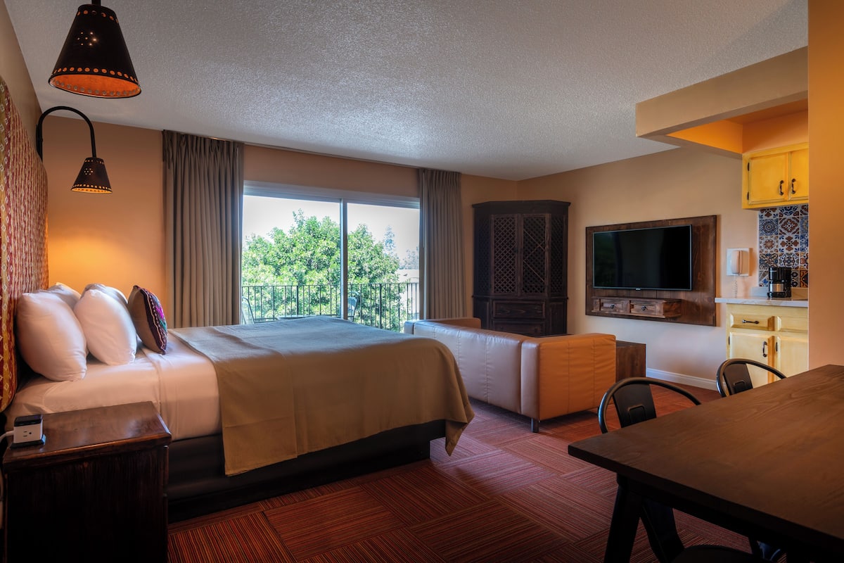 A hotel room with a large bed, a desk, a dining table, a TV, and a balcony with a view of trees.